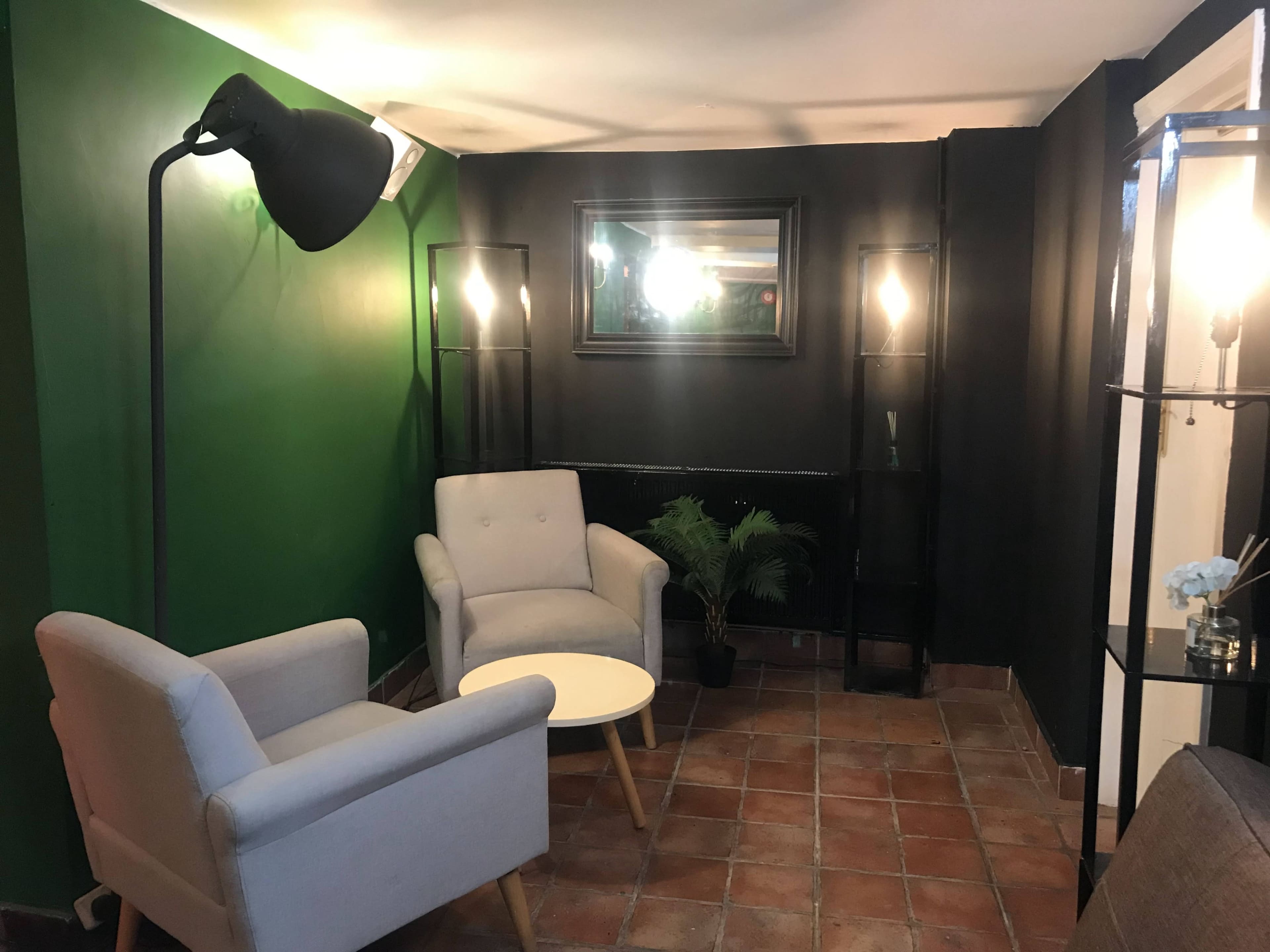 A cozy seating area with two light gray armchairs, a small circular table, and decorative floor lamps against a dark green and black wall.
