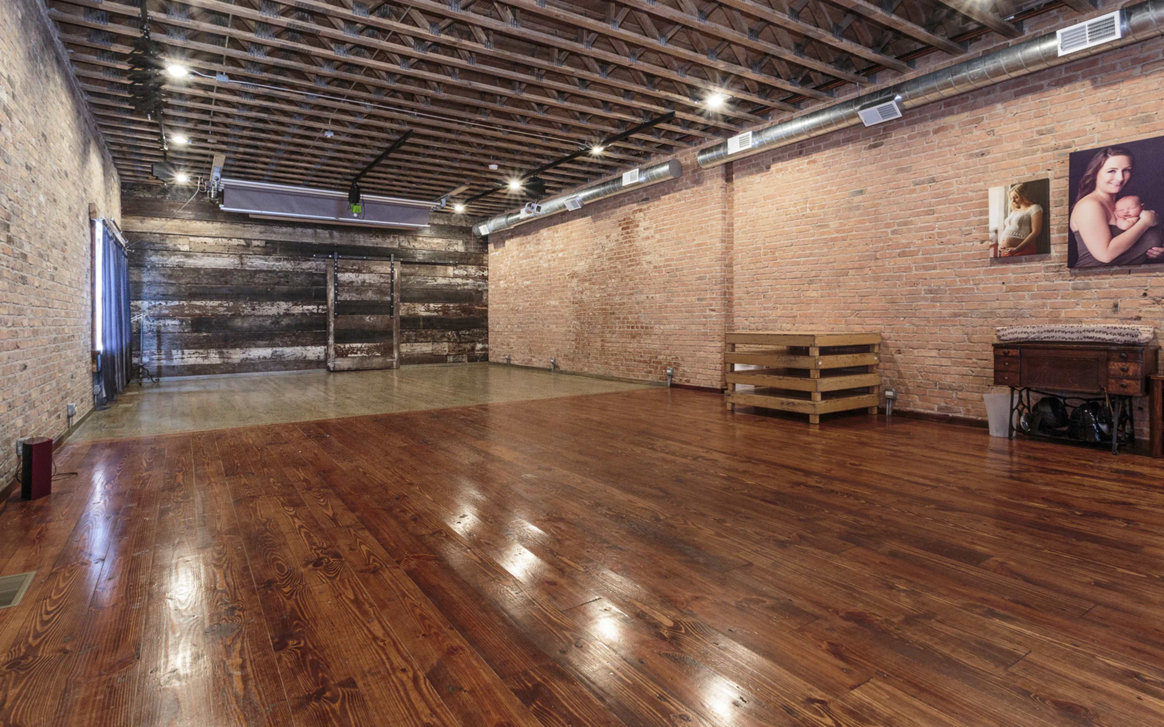 A spacious, empty studio with exposed brick walls, wooden flooring, and large windows, featuring a rustic ambiance.
