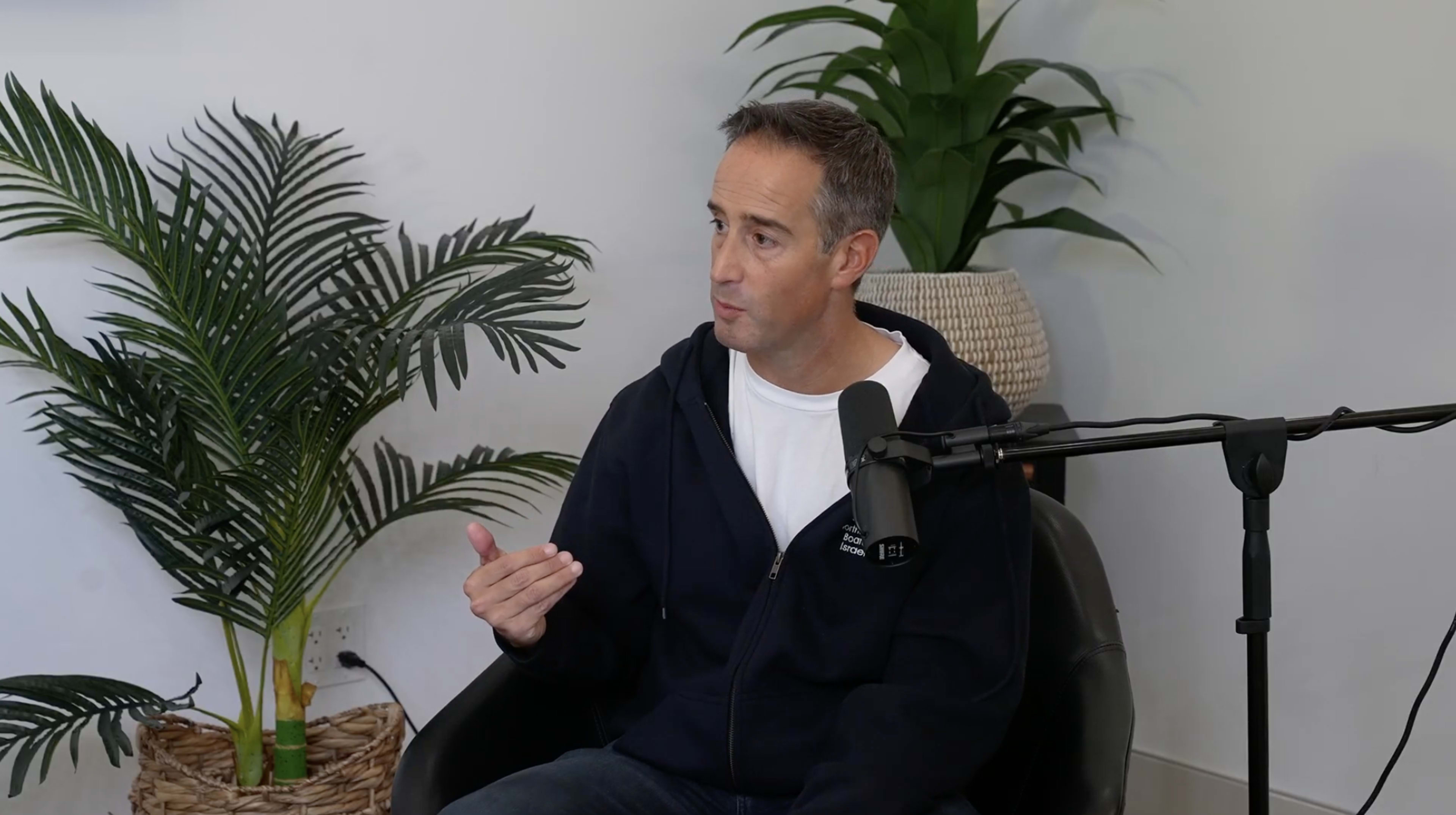 A person is seated in a black chair, speaking into a microphone while surrounded by potted plants and a woven basket.
