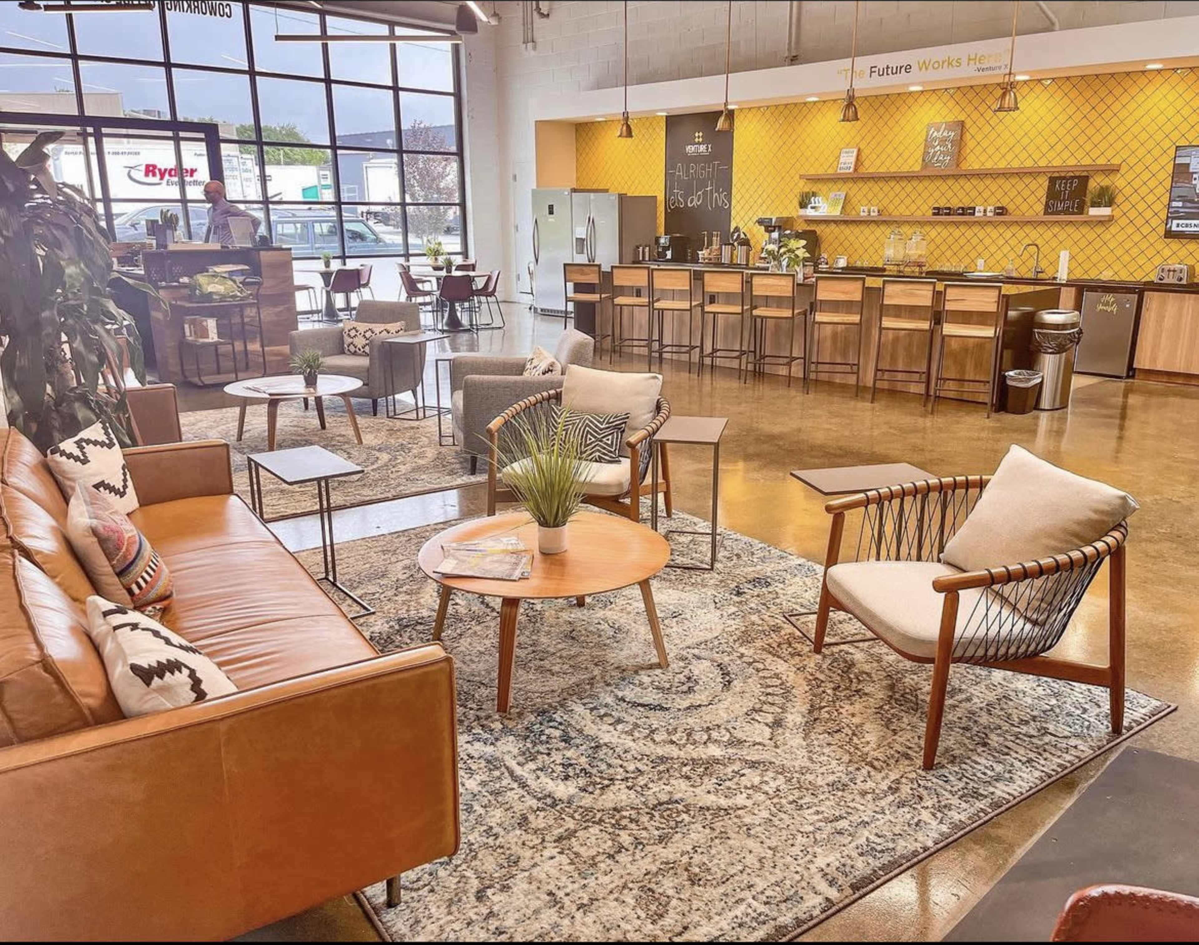 A modern lounge area featuring brown leather sofas, mid-century style chairs, a circular coffee table, and a large patterned rug, complemented by a barista station in the background.