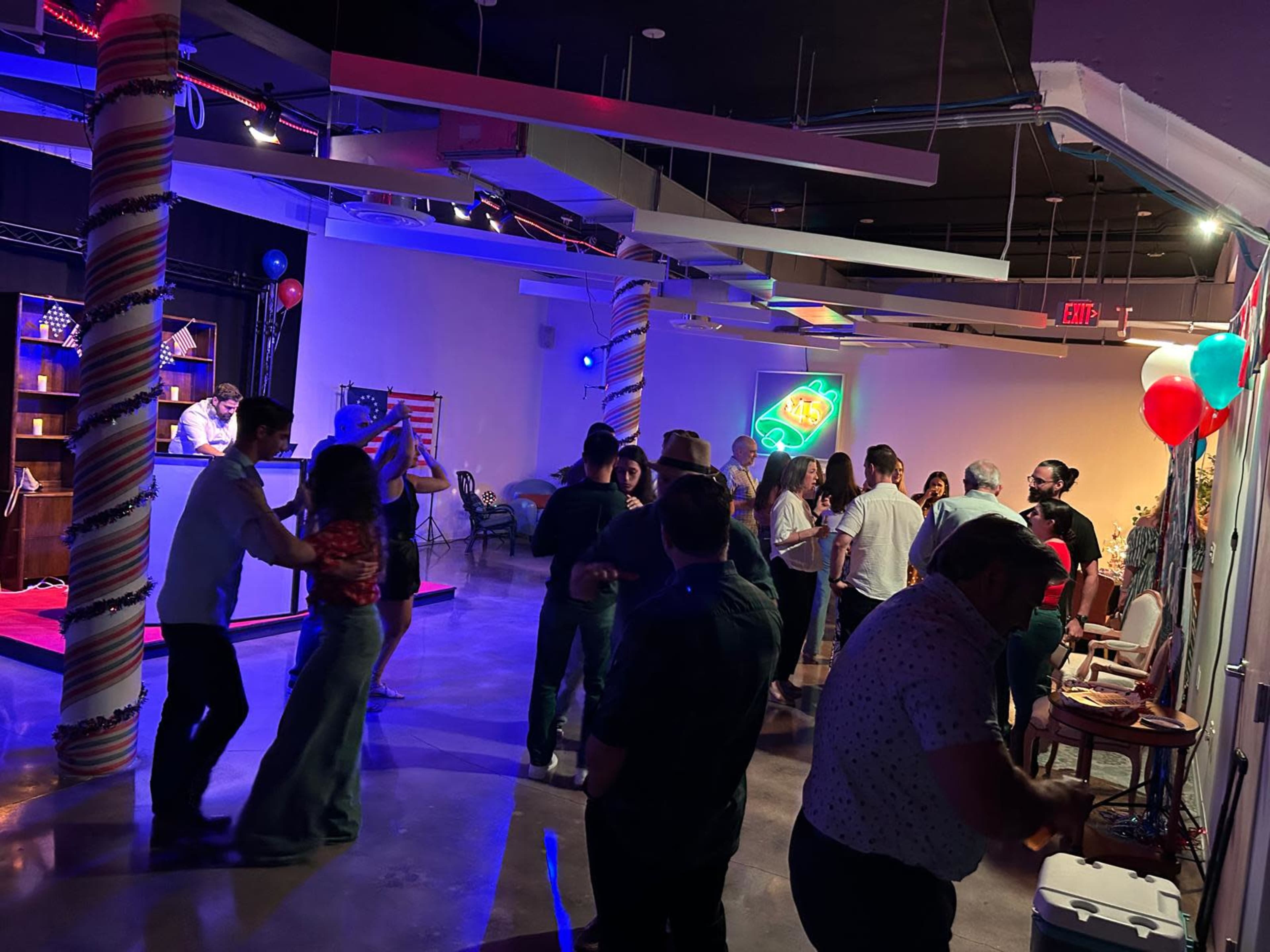 People are dancing and socializing at a lively indoor event with colorful decorations and a DJ in the background.