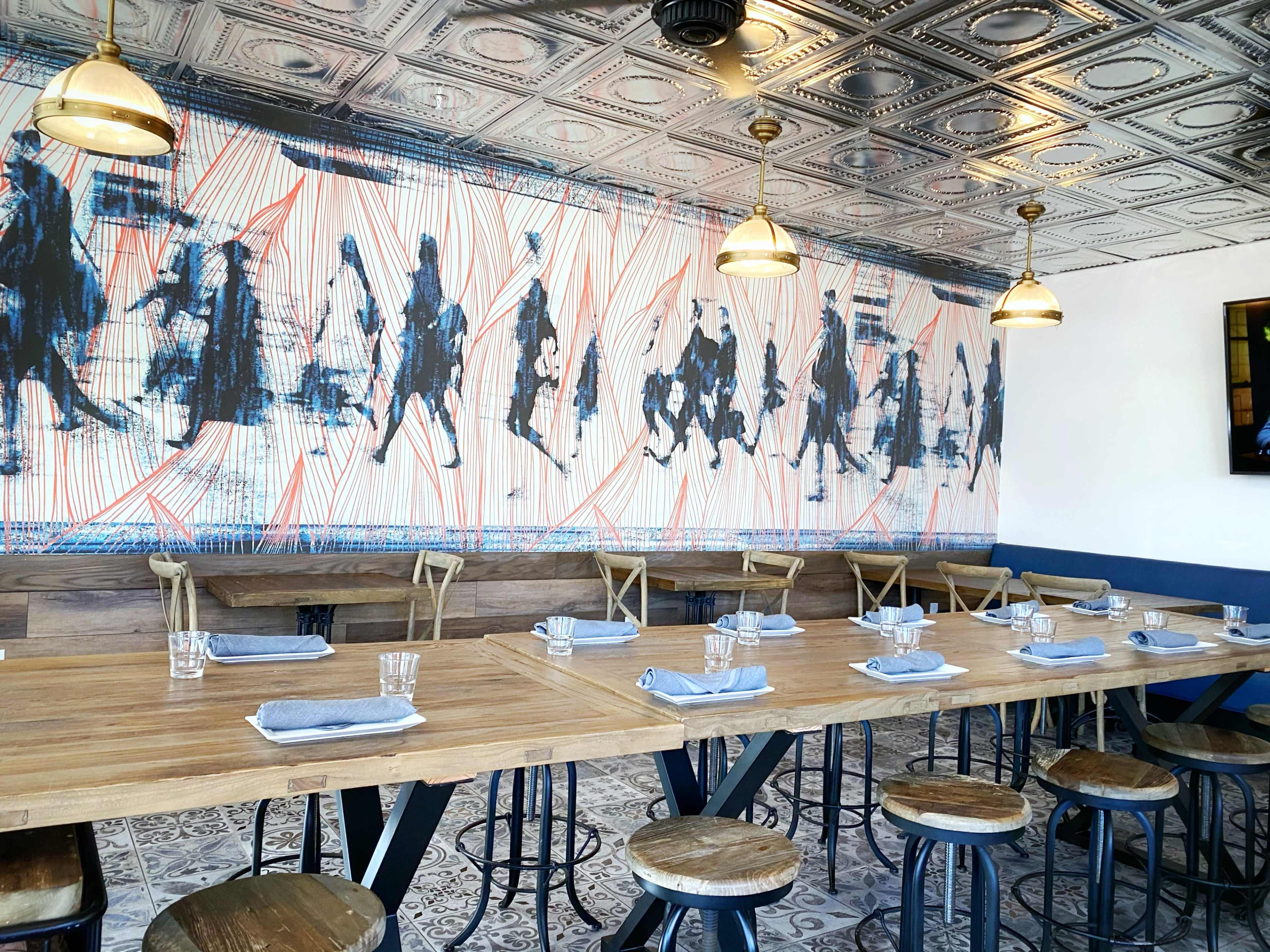 A dining area features a long wooden table set with glasses and napkins, surrounded by stools, and is adorned with a mural of silhouetted figures on the wall.