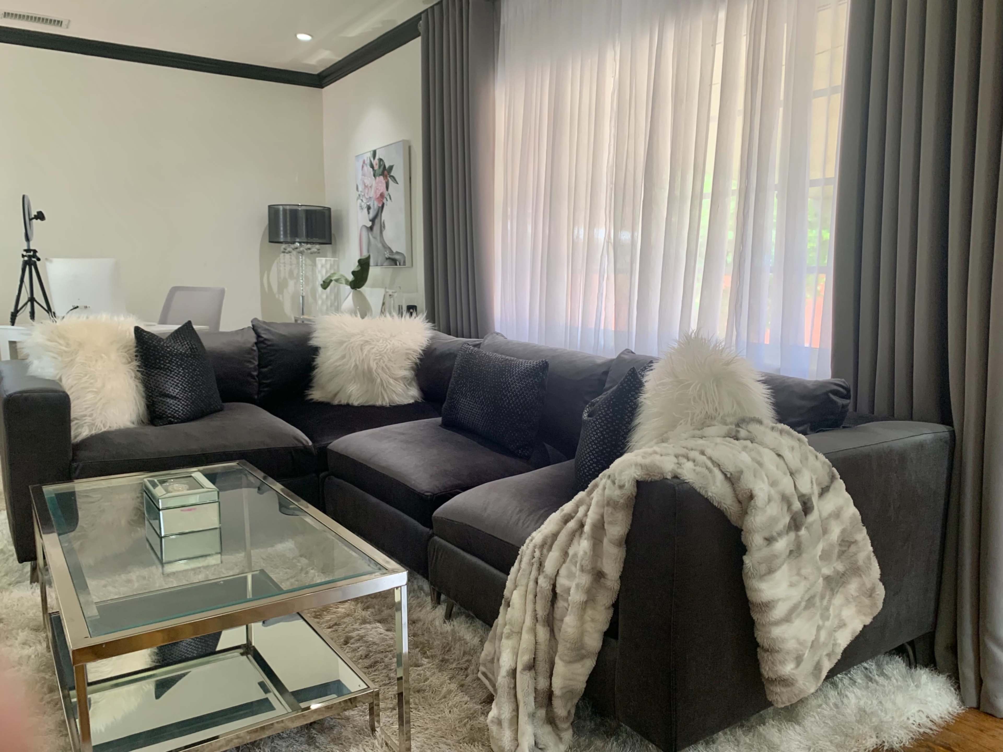 A modern living room featuring a dark sectional sofa adorned with fluffy pillows and a cozy throw, a glass coffee table, and large windows with sheer curtains.
