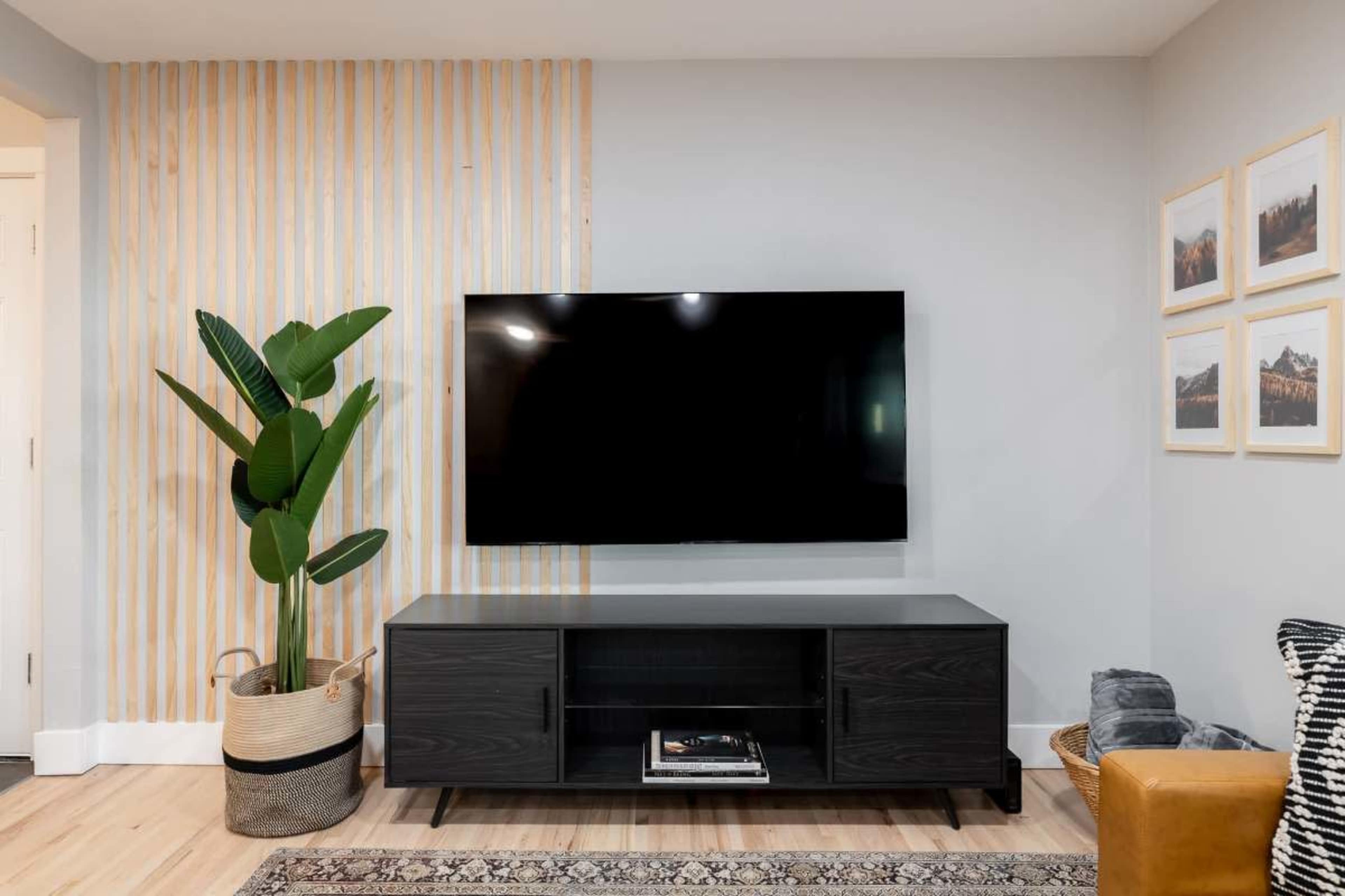 The image shows a modern living room with a large television mounted on the wall above a dark wooden media console, accompanied by a potted plant and framed photographs on the adjacent wall.