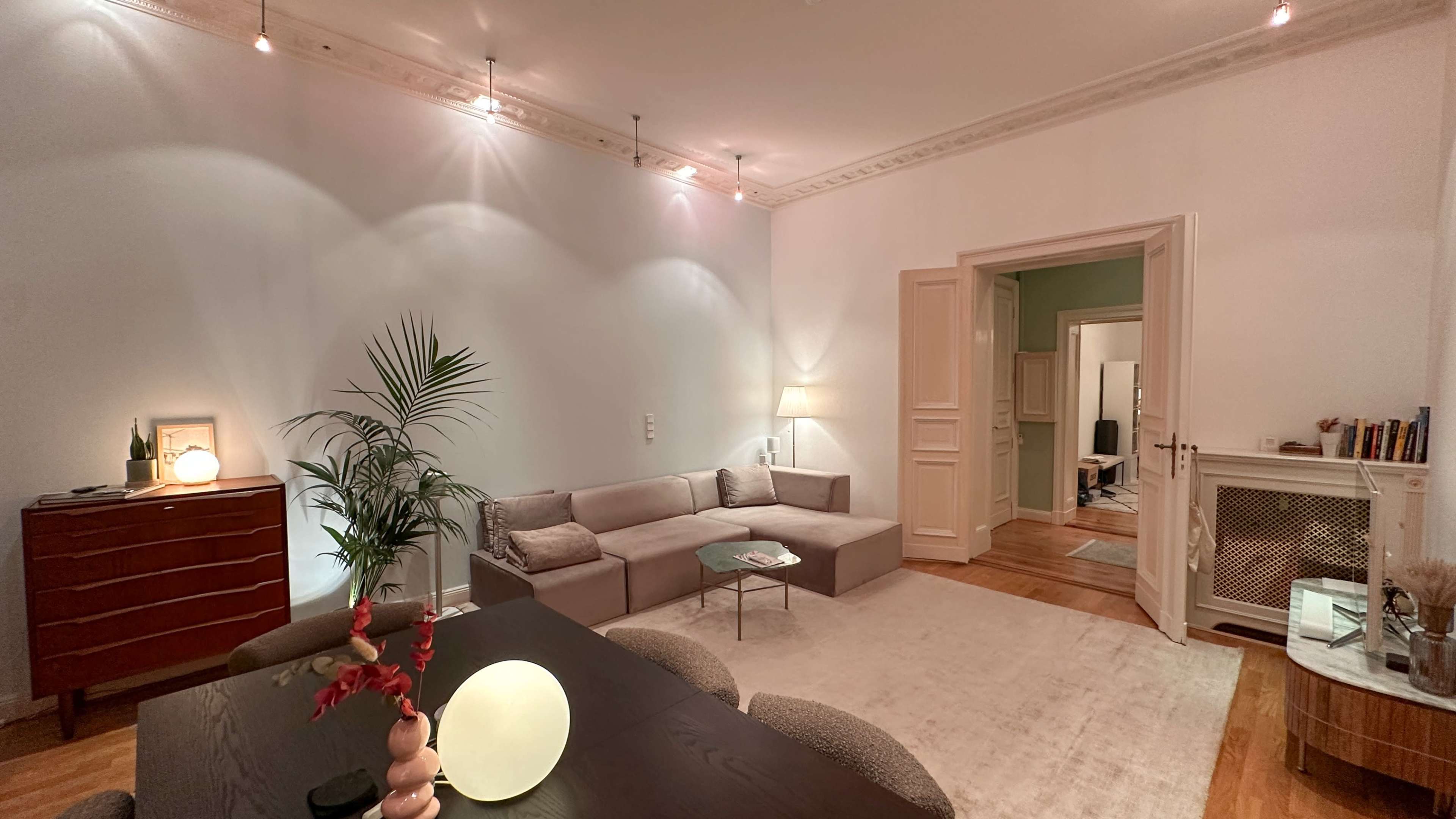 The image shows a well-lit living room featuring a gray sectional sofa, a round light, and a wooden dining table with upholstered chairs.