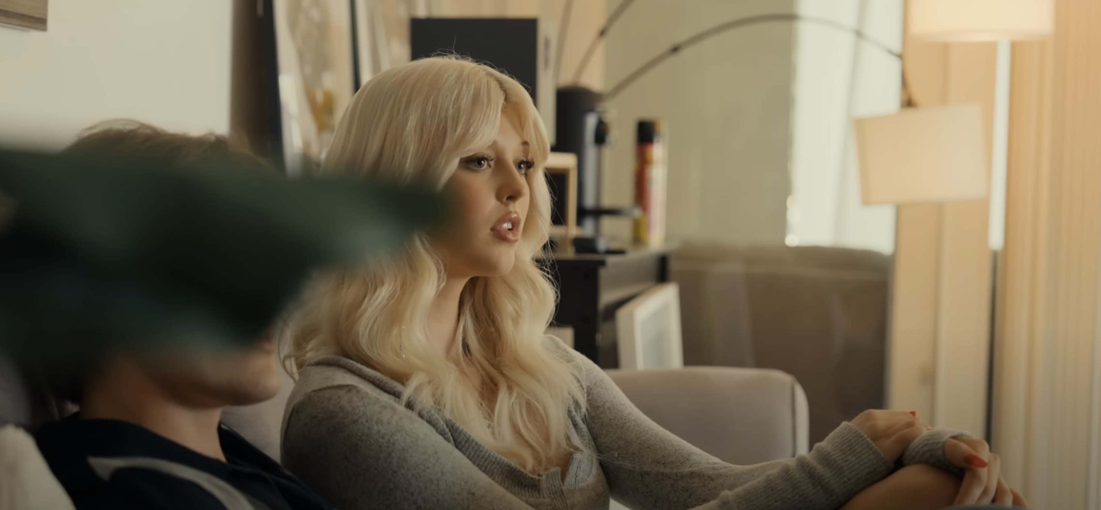 A woman with long blonde hair sits on a couch, speaking to a person partially obscured by a plant in a well-lit living room.