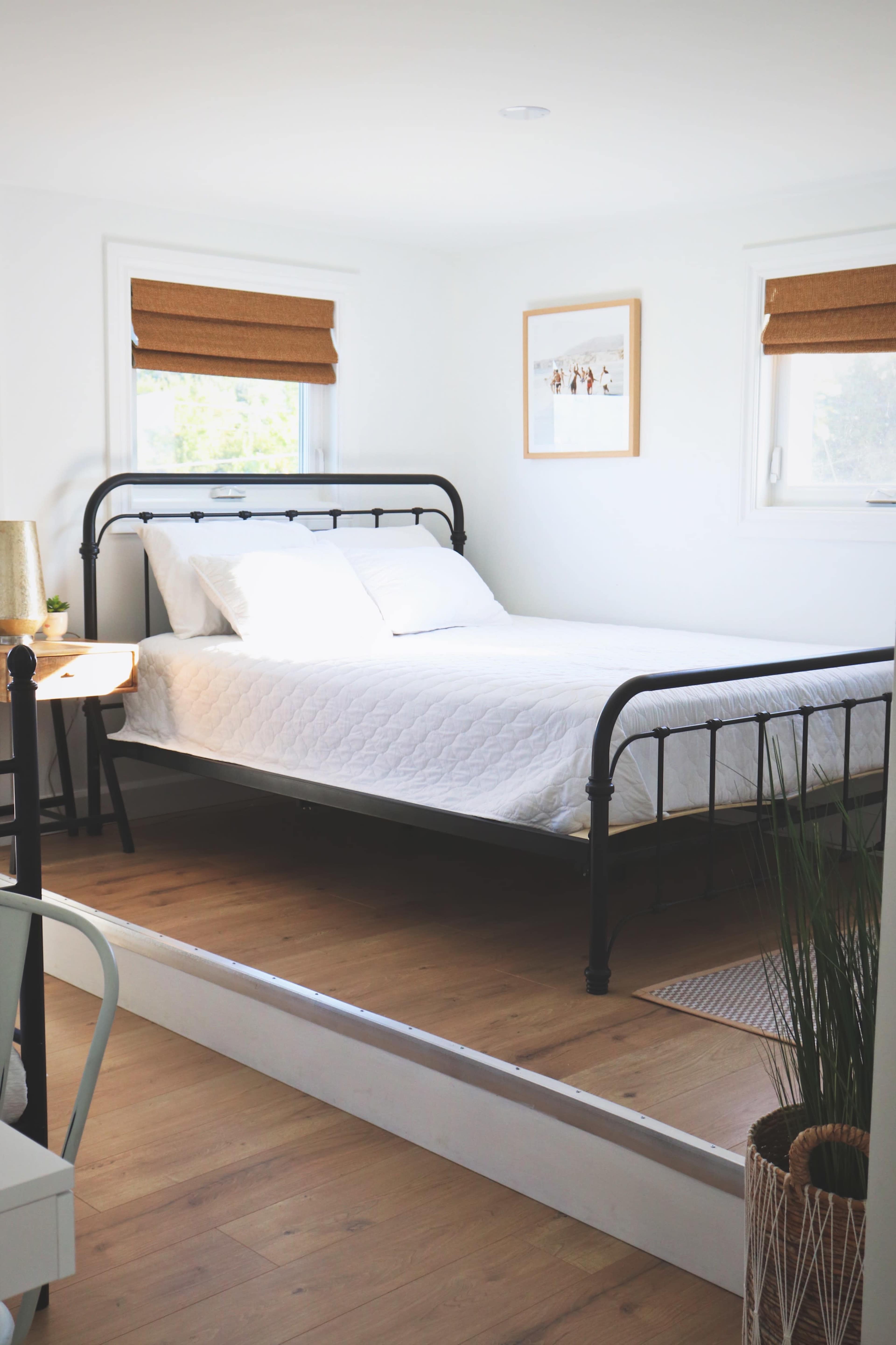 A simple bedroom features a metal bed frame with a white quilt and a small side table beside it, illuminated by natural light through window shades.