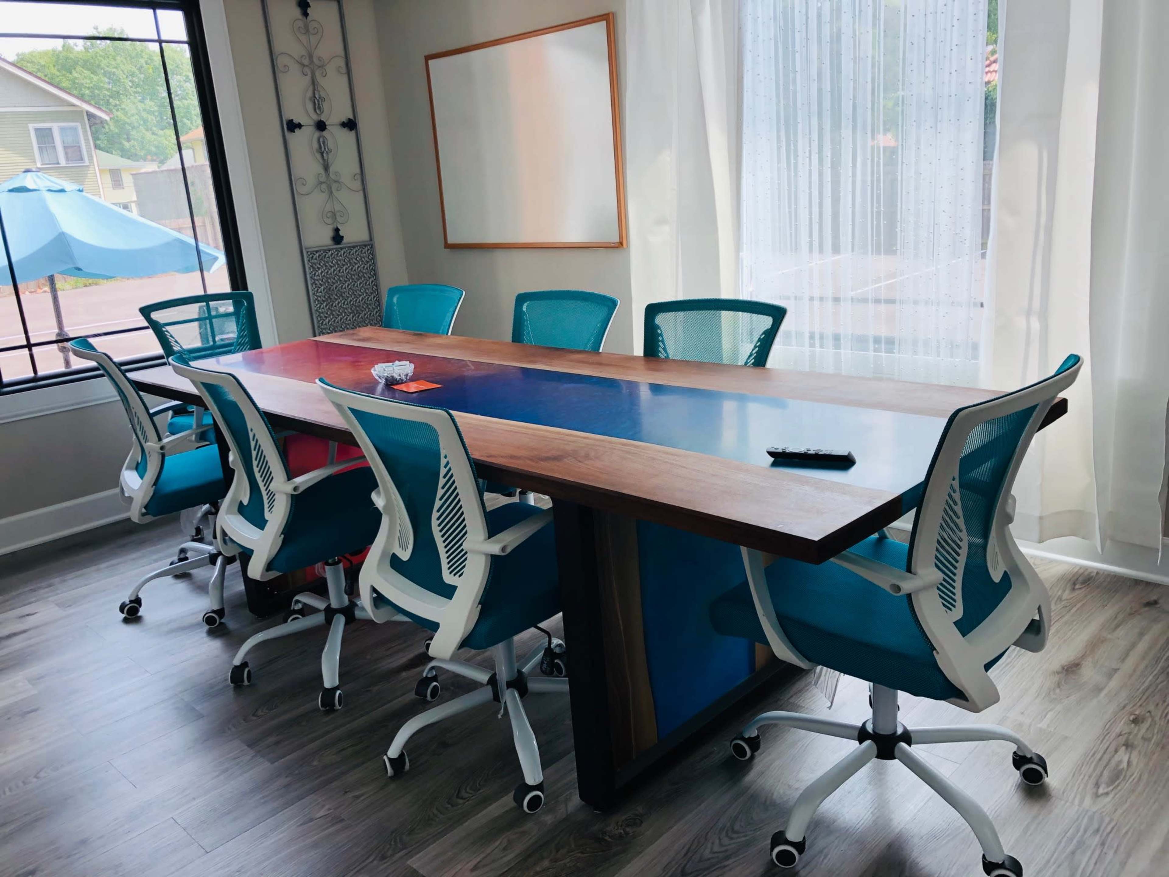The image shows a conference room with a large wooden table and eight blue and white office chairs arranged around it, illuminated by natural light from a window.