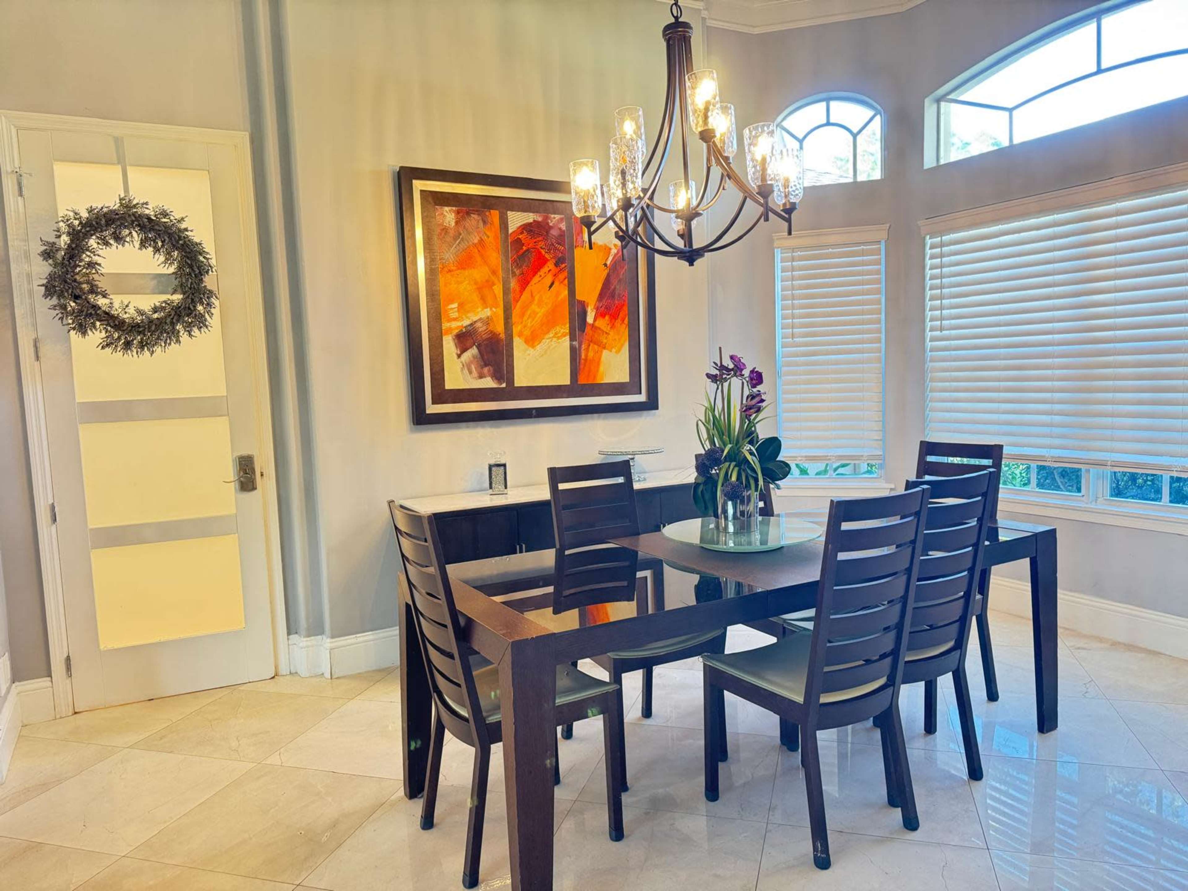 A dining area features a wooden table surrounded by six chairs, a colorful abstract painting on the wall, and a chandelier above.