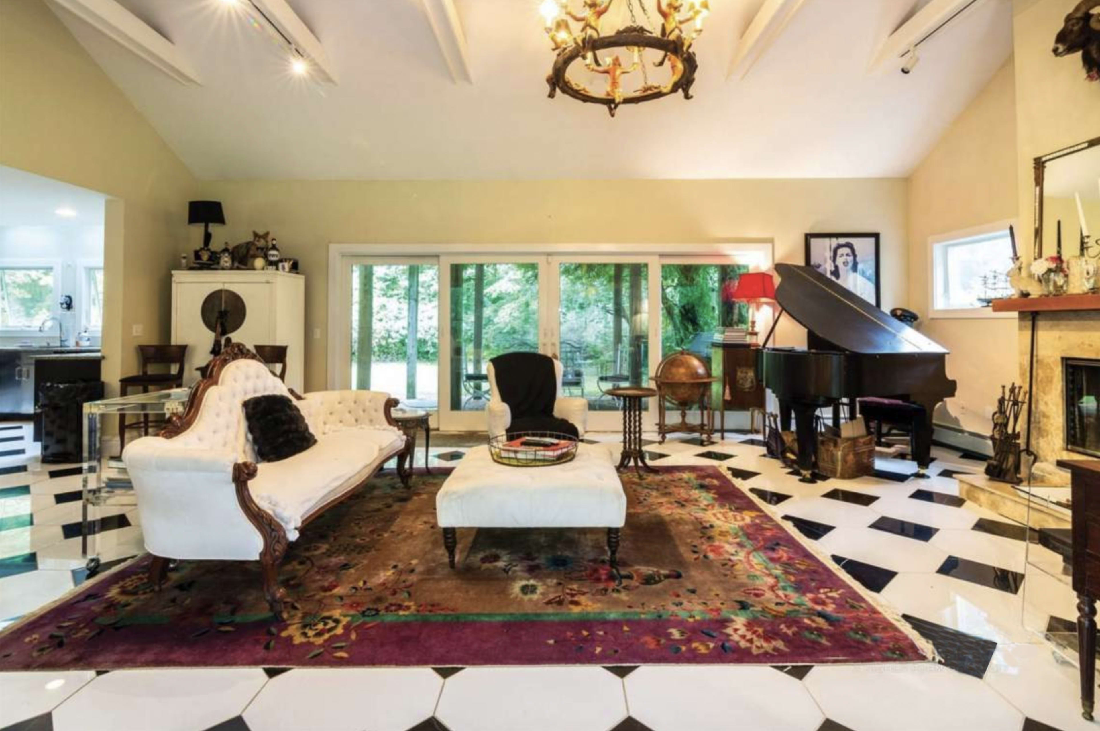 A spacious living room with a chandelier, a piano, a white sofa, a decorative rug, and large windows overlooking a garden.