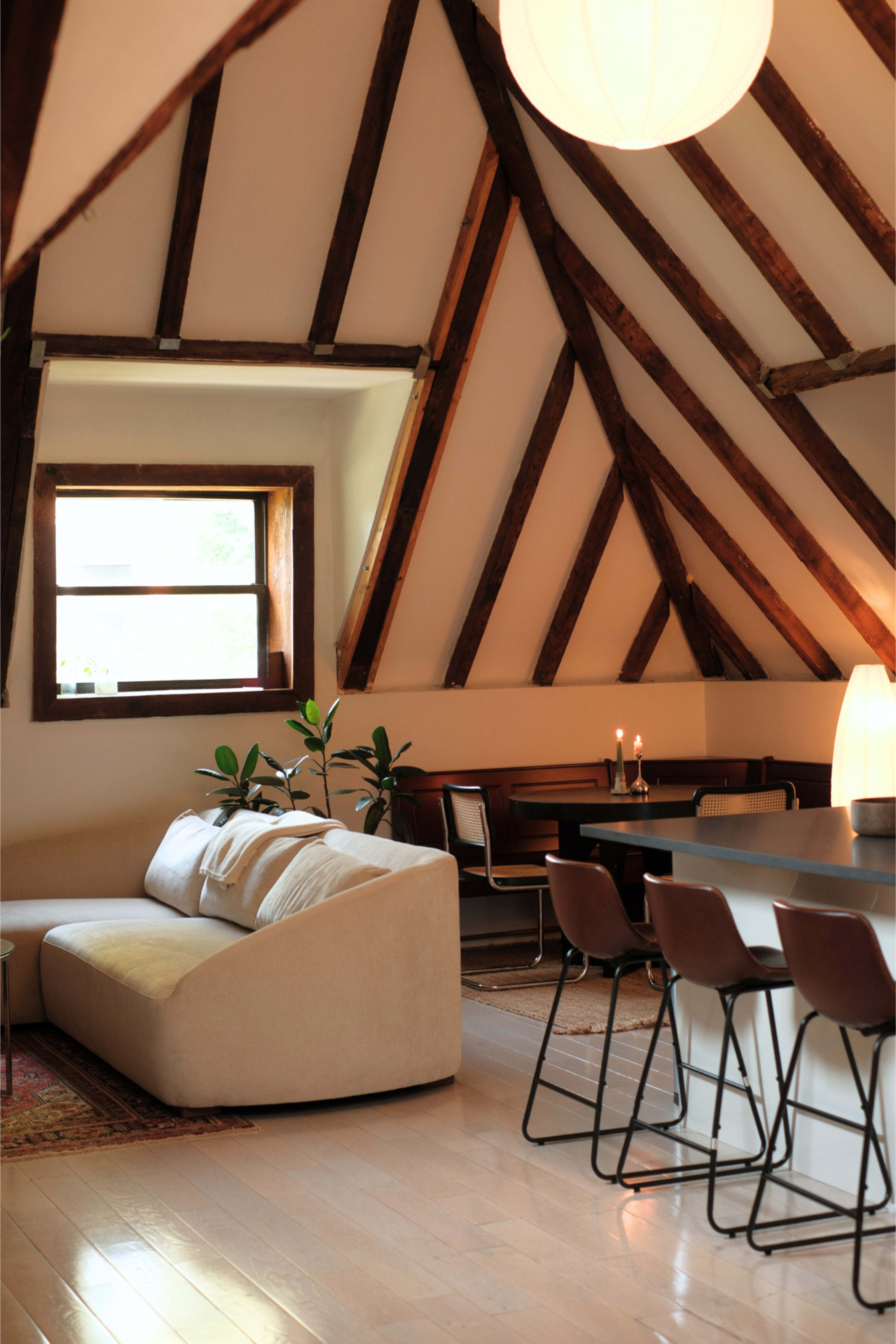 The image shows a modern living space with exposed wooden beams, a beige sofa, a dining table with chairs, and soft lighting.