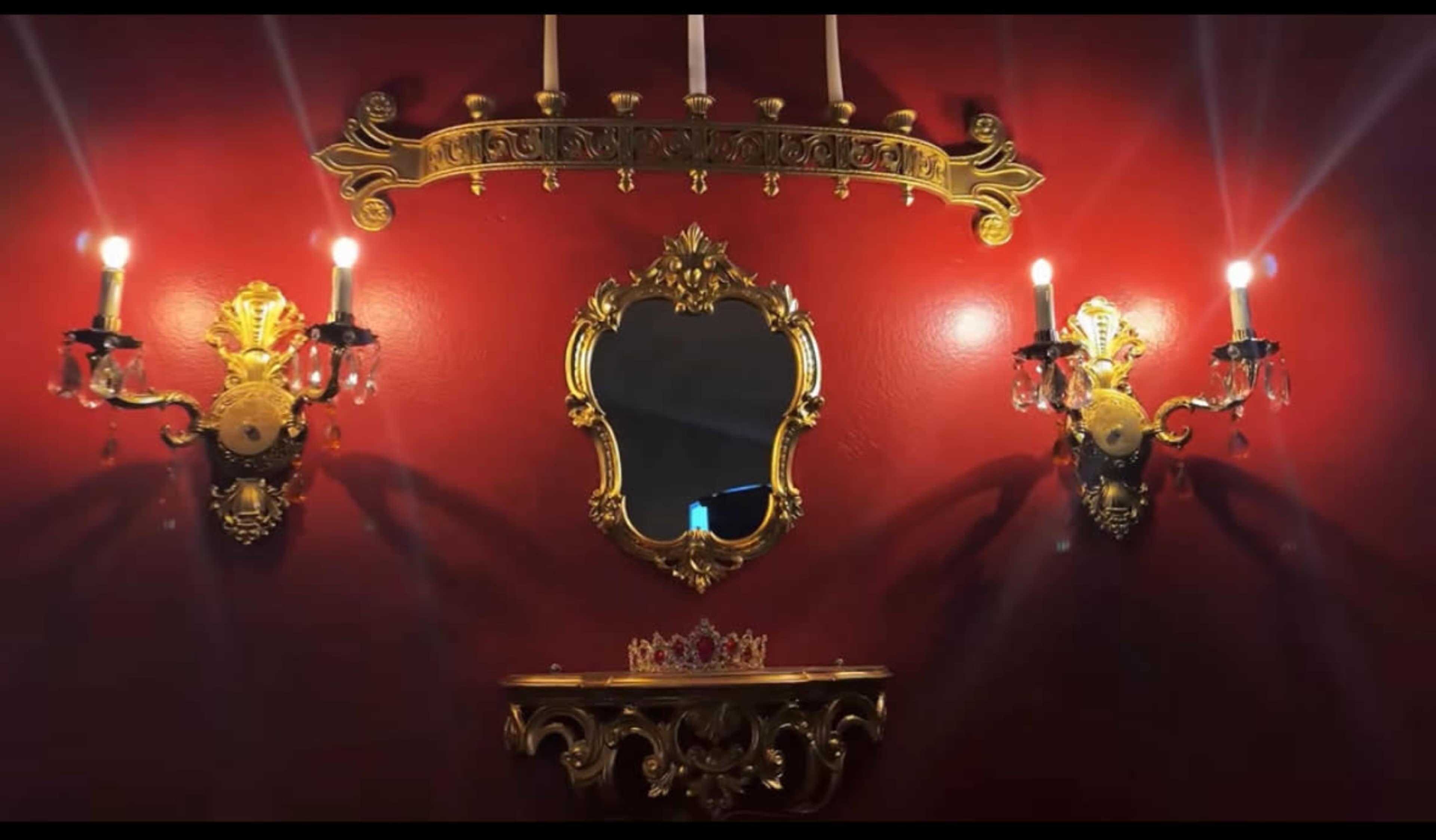 The image shows a richly decorated red wall adorned with ornate gold sconces and a central mirror, complemented by a decorative shelf and a crown.