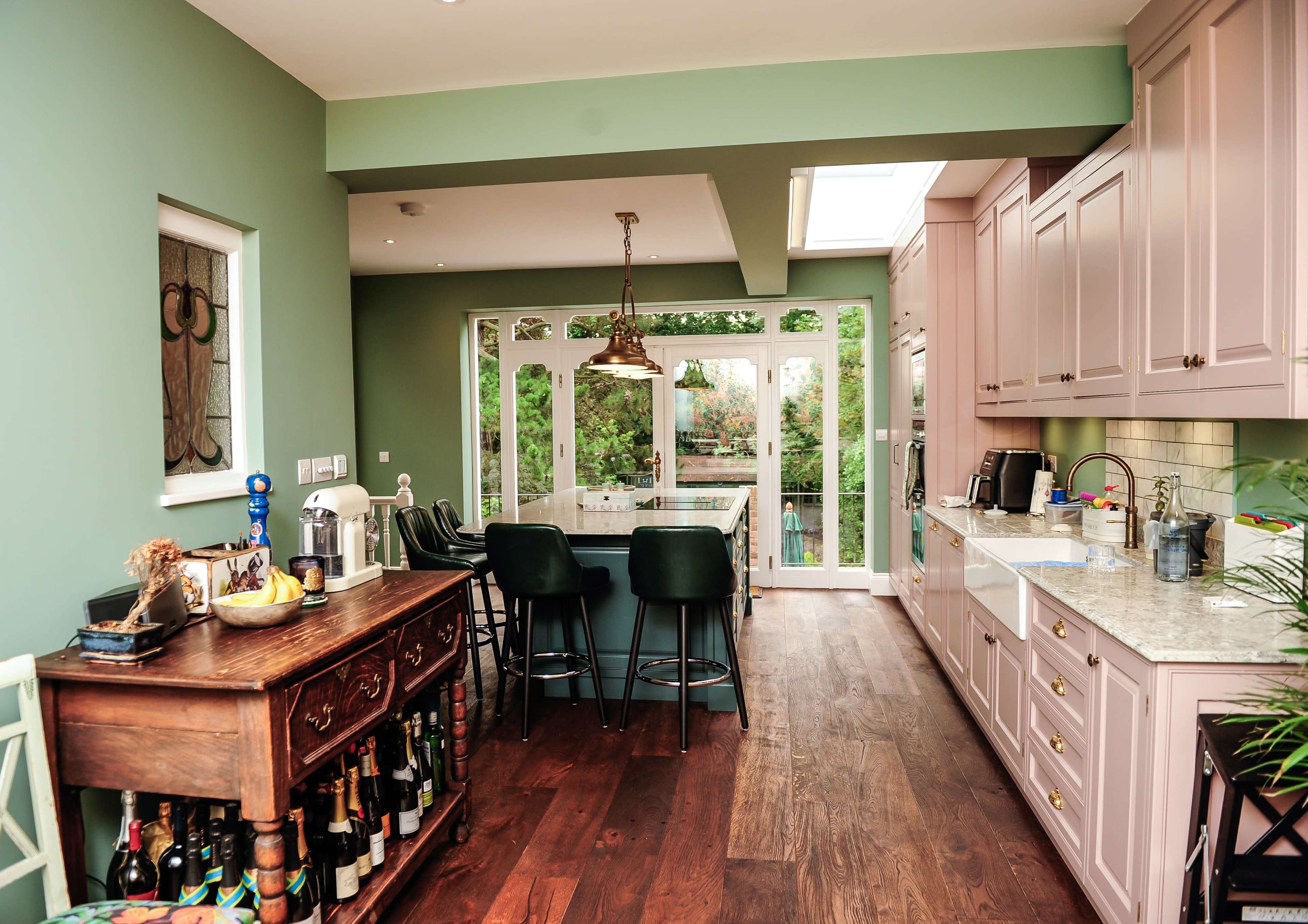 A modern kitchen features light green walls, pink cabinets, a marble countertop, and a view of a garden through large glass doors.