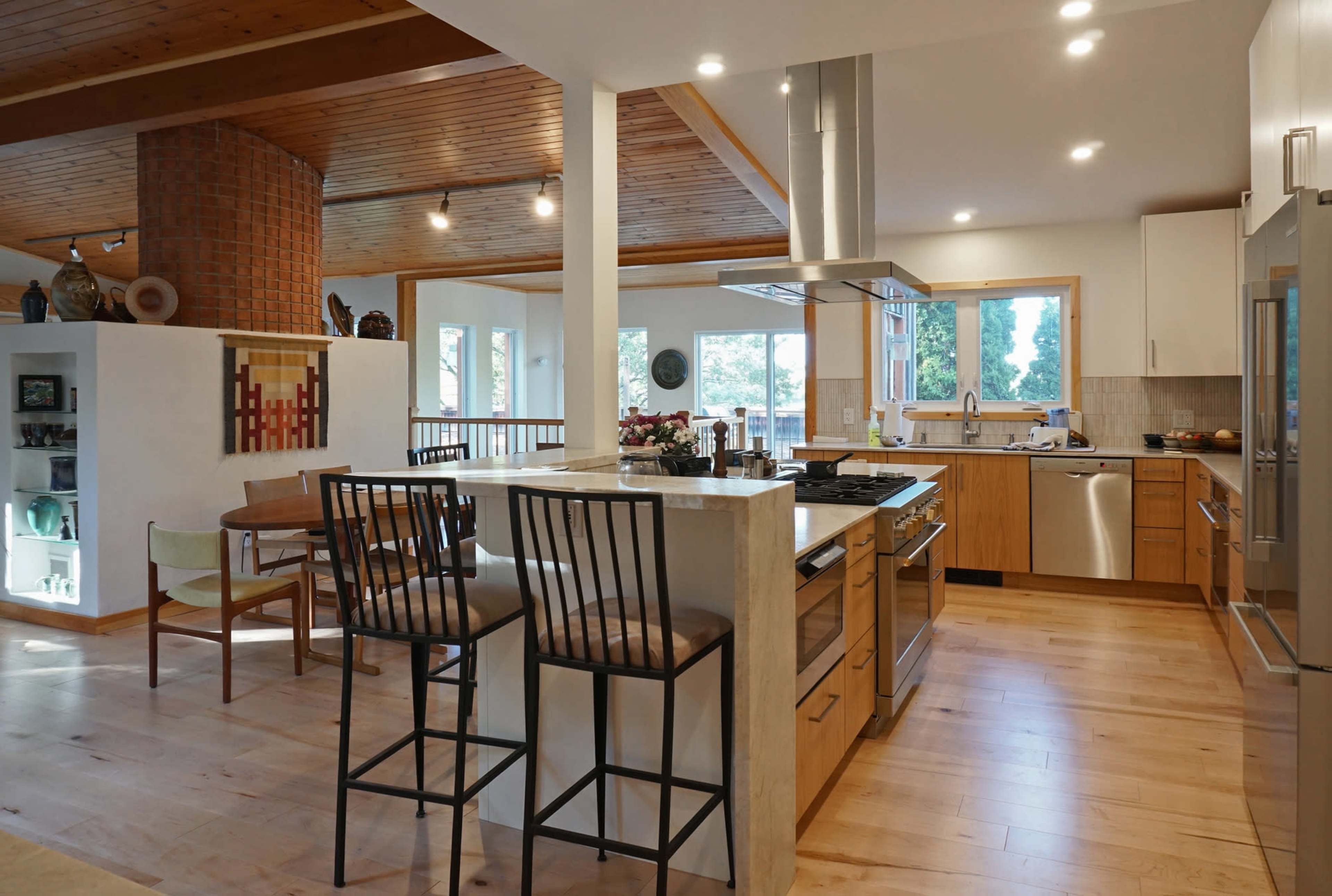 The image shows a modern kitchen with wooden cabinetry, a large island with bar stools, and ample natural light coming through multiple windows.
