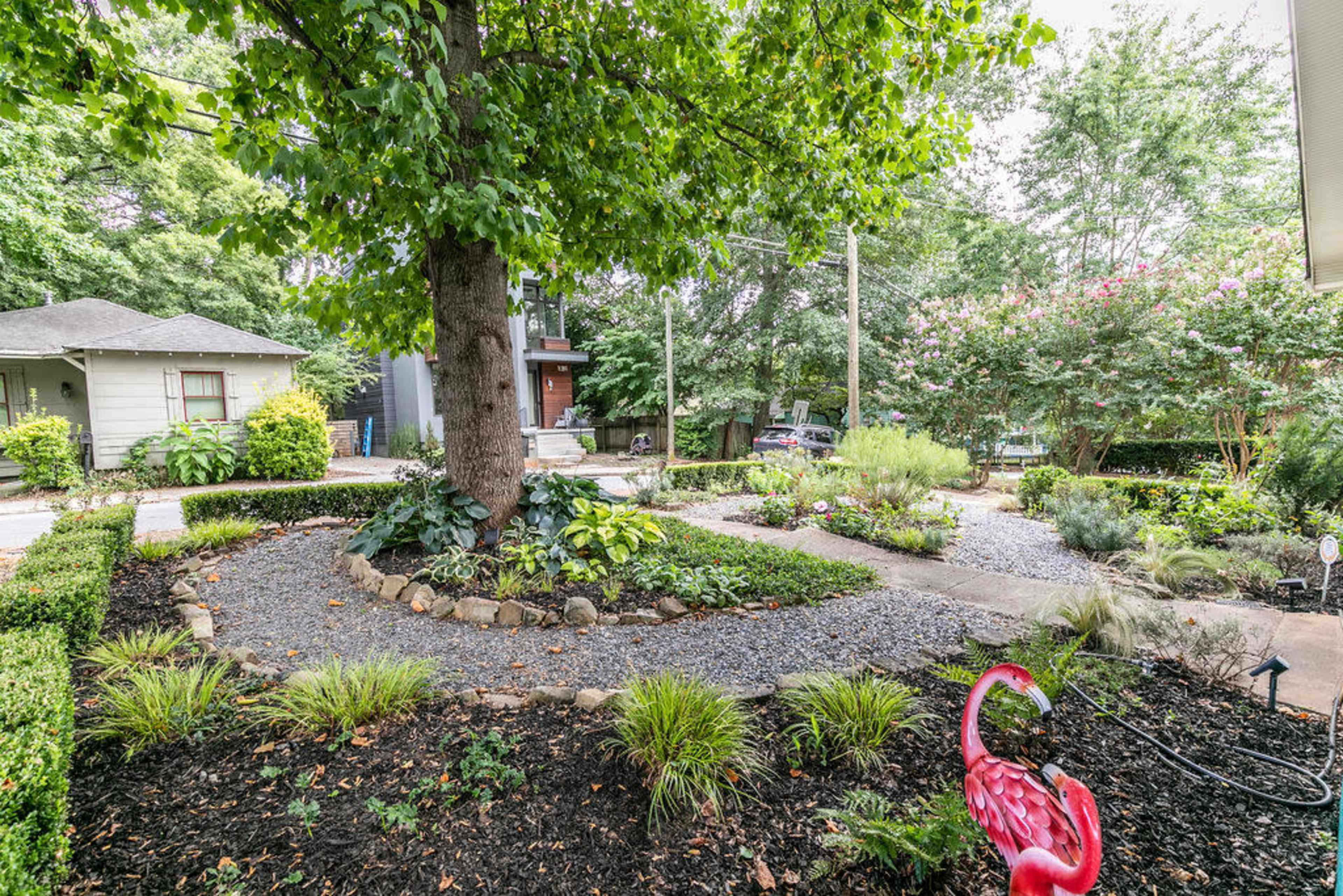 A landscaped garden features a large tree at its center, surrounded by gravel pathways and various plants, with two flamingo sculptures in the foreground.