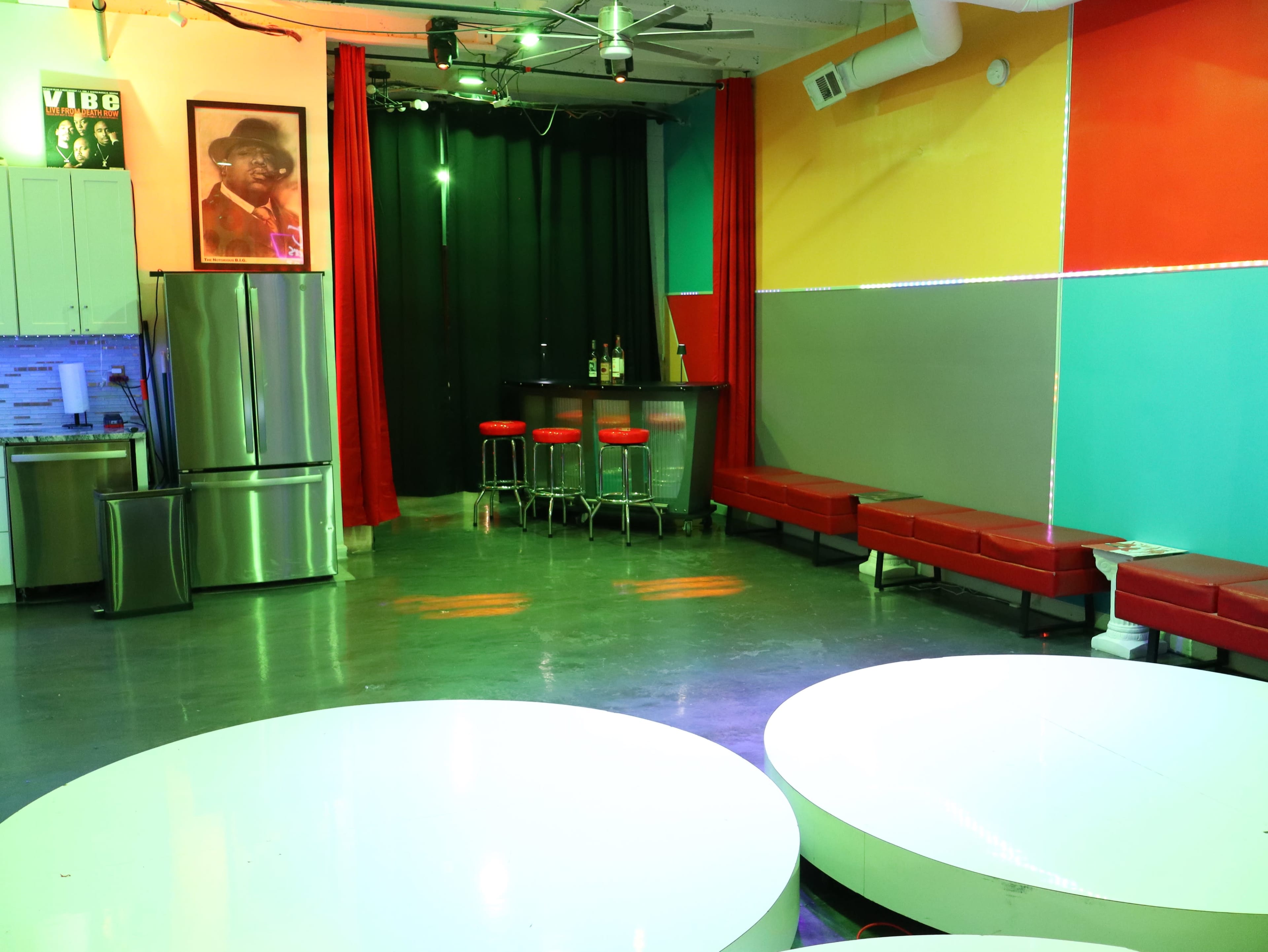 The image shows a colorful interior space with red benches, round white tables, a bar area, and a large wall-mounted photo.
