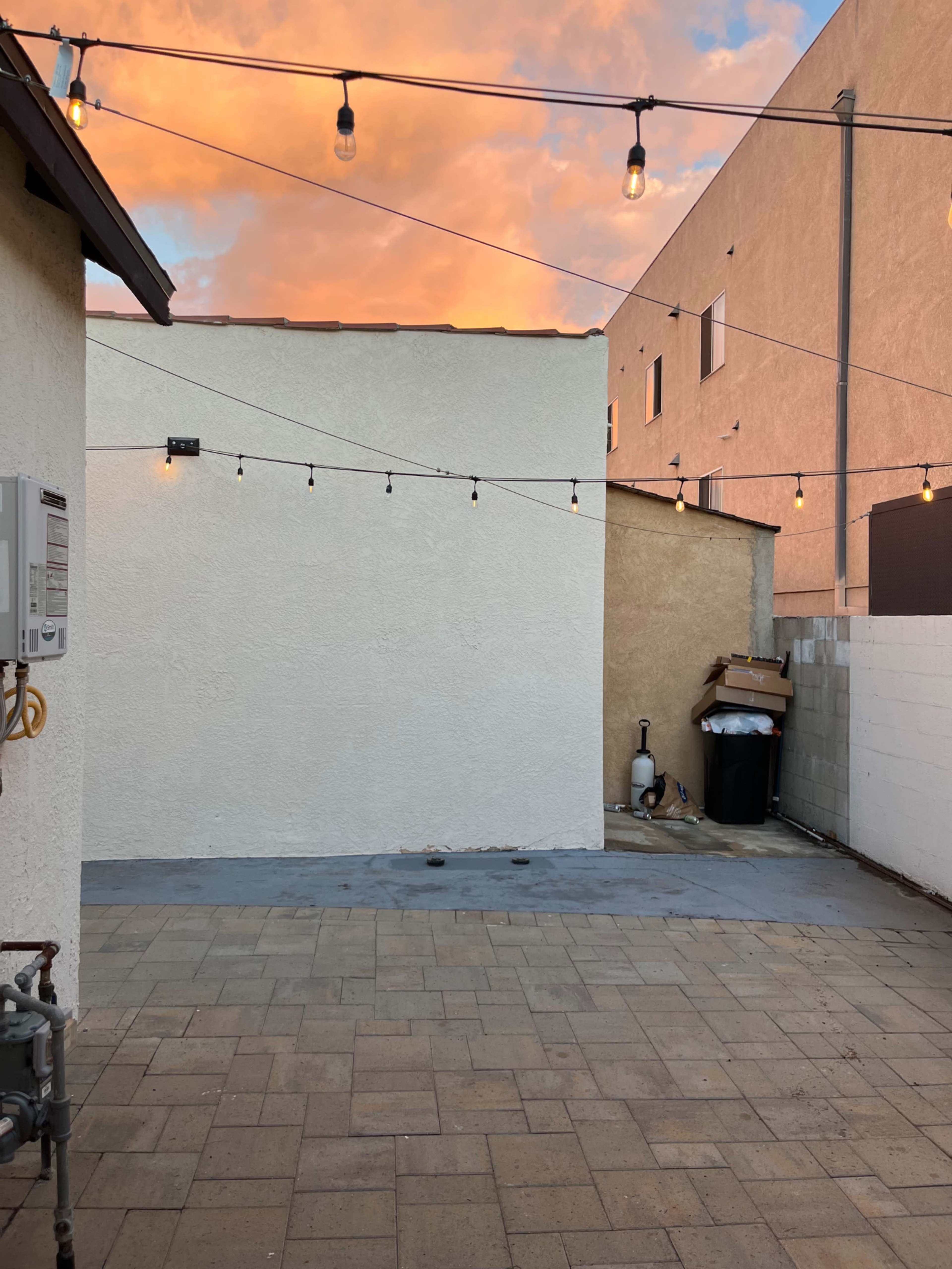 A small outdoor area with paved flooring, a white wall, and string lights overhead, set against a colorful sky at sunset.