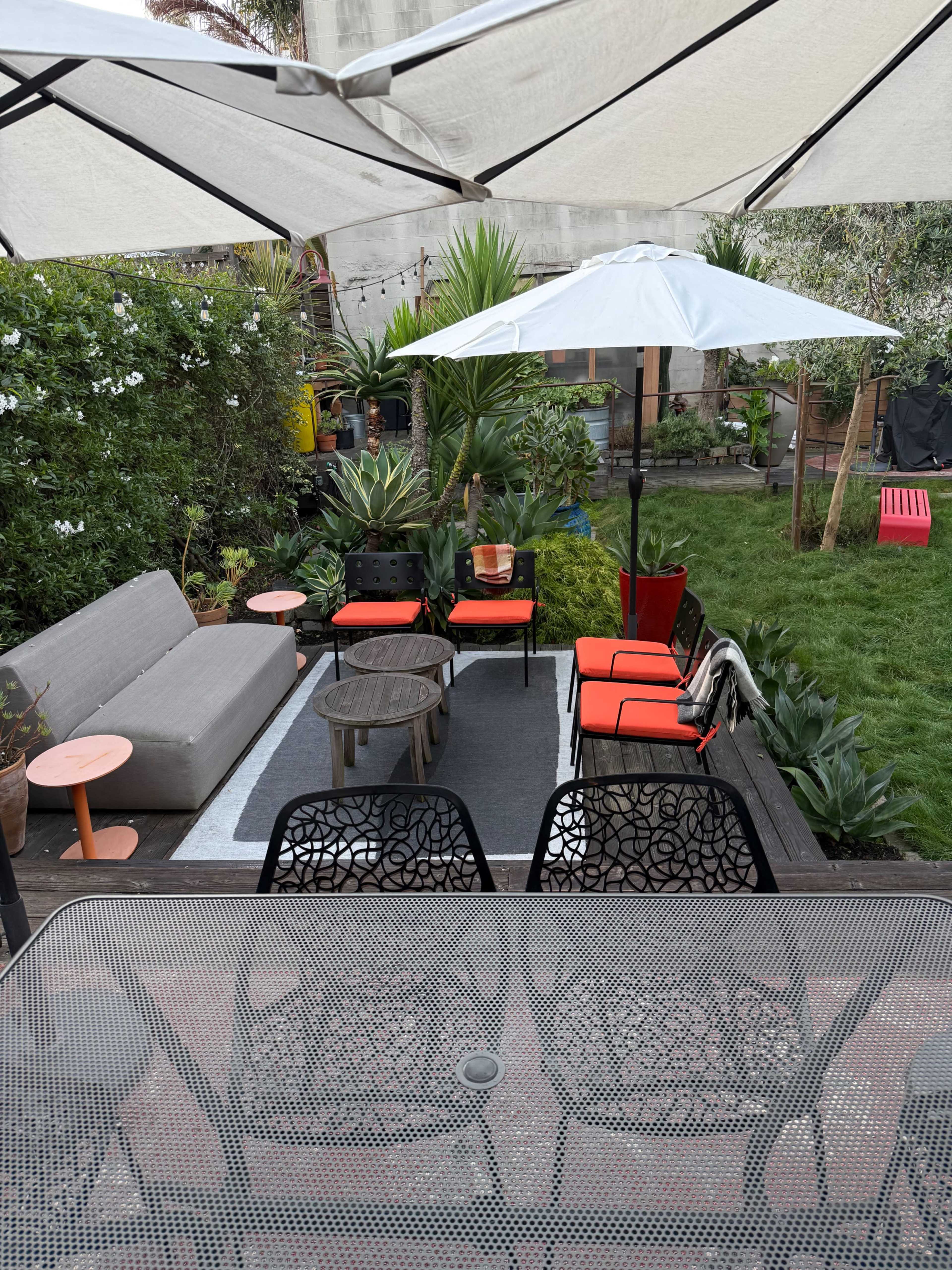 The image shows a small outdoor garden area with a seating arrangement that includes a gray couch, two red chairs, and a round table, surrounded by plants and shaded by large umbrellas.