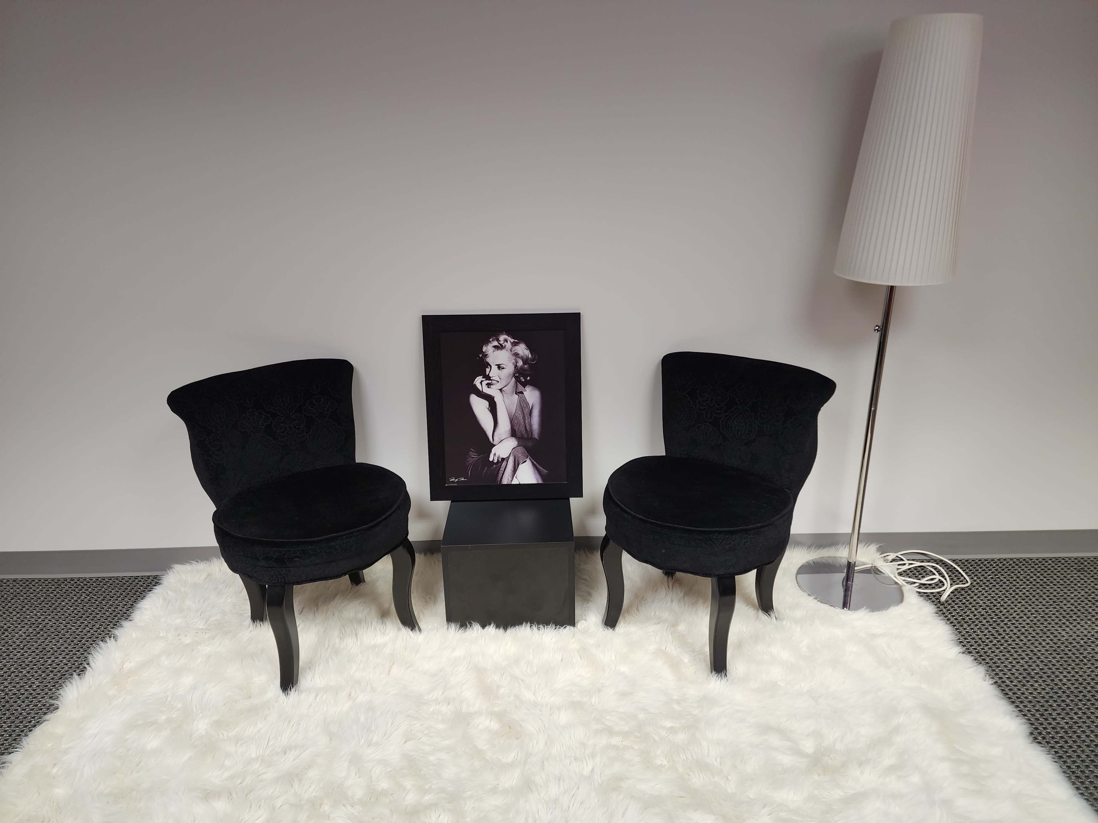 Two black upholstered chairs facing a black square table, with a framed black-and-white portrait and a tall white lamp in a minimalist room setting on a fluffy white rug.