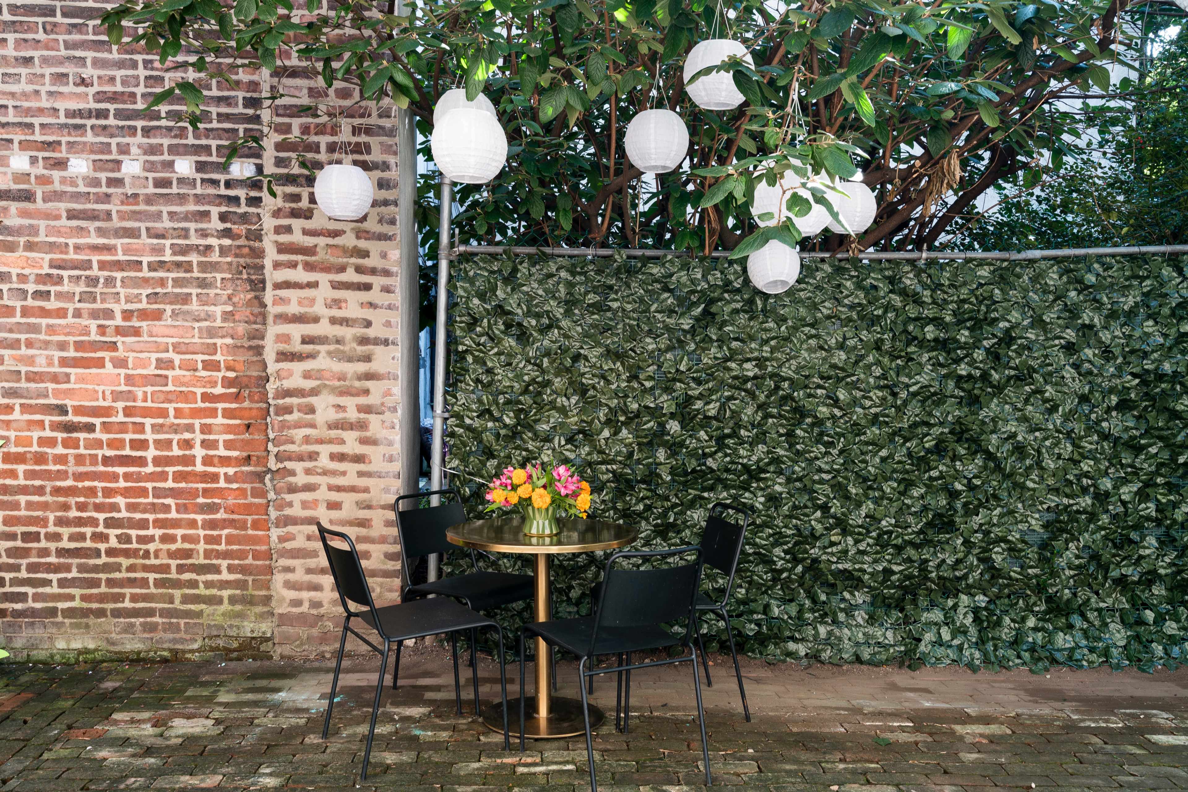 A small round table with a flower arrangement is surrounded by four black chairs beneath hanging lanterns, next to a brick wall and a leafy green backdrop.