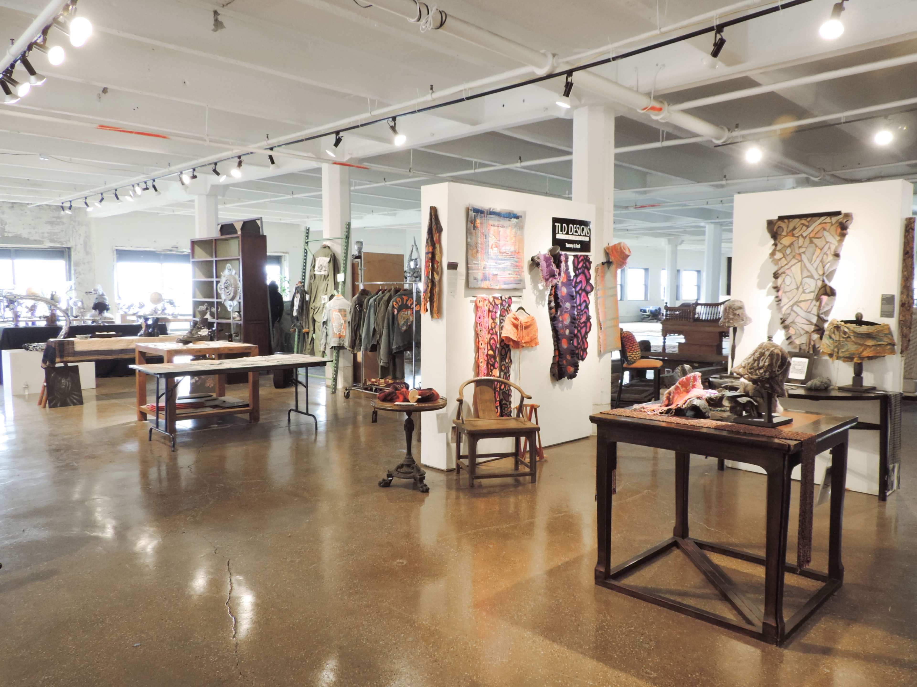 The image shows an art gallery with various display tables and walls featuring textiles and artistic objects in a spacious, well-lit environment.