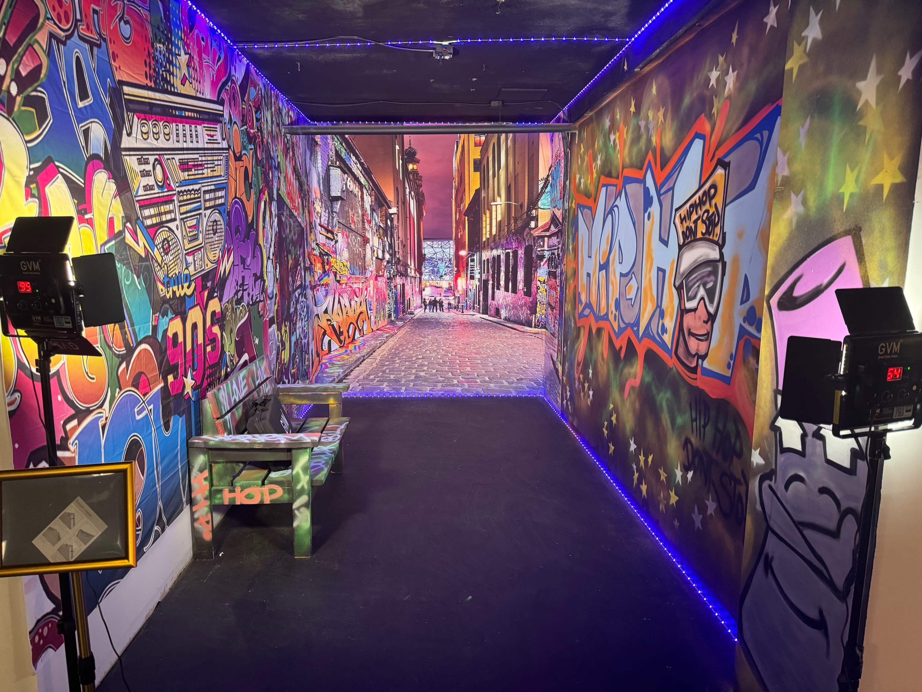 The image shows a graffiti-covered corridor that leads to a scenic street view, illuminated by colorful lights.