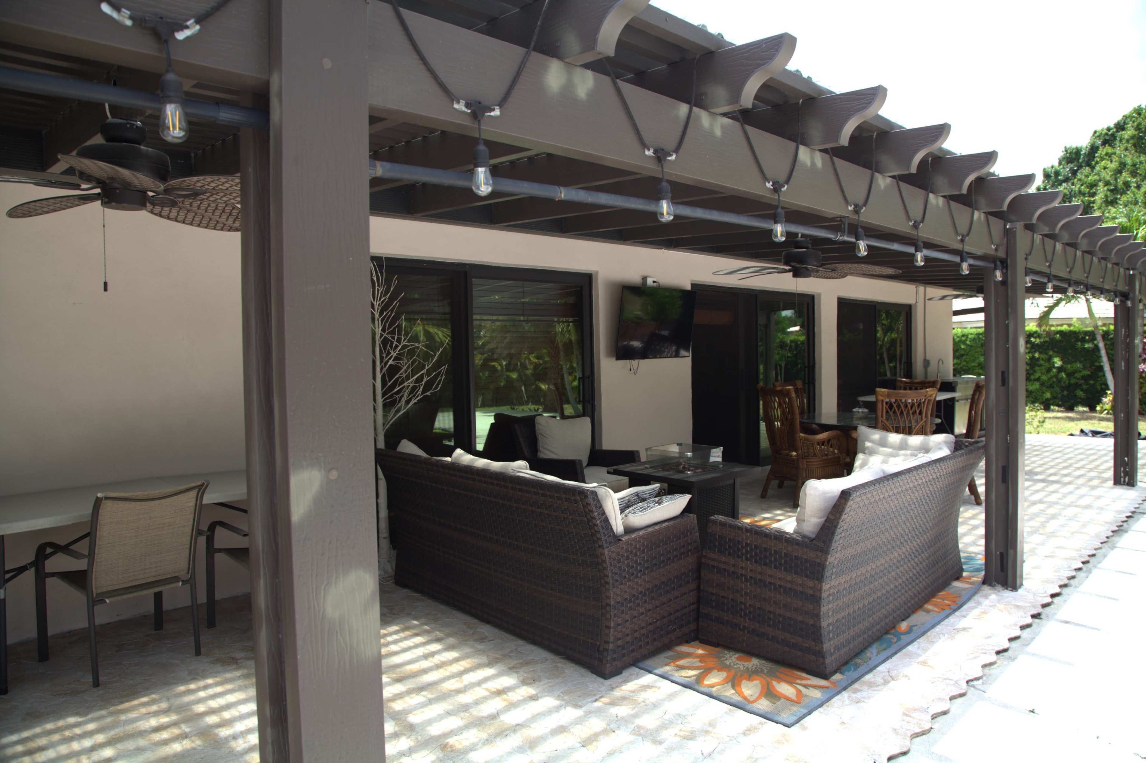 The image shows a covered patio with outdoor furniture, including a seating area and dining table, surrounded by greenery.