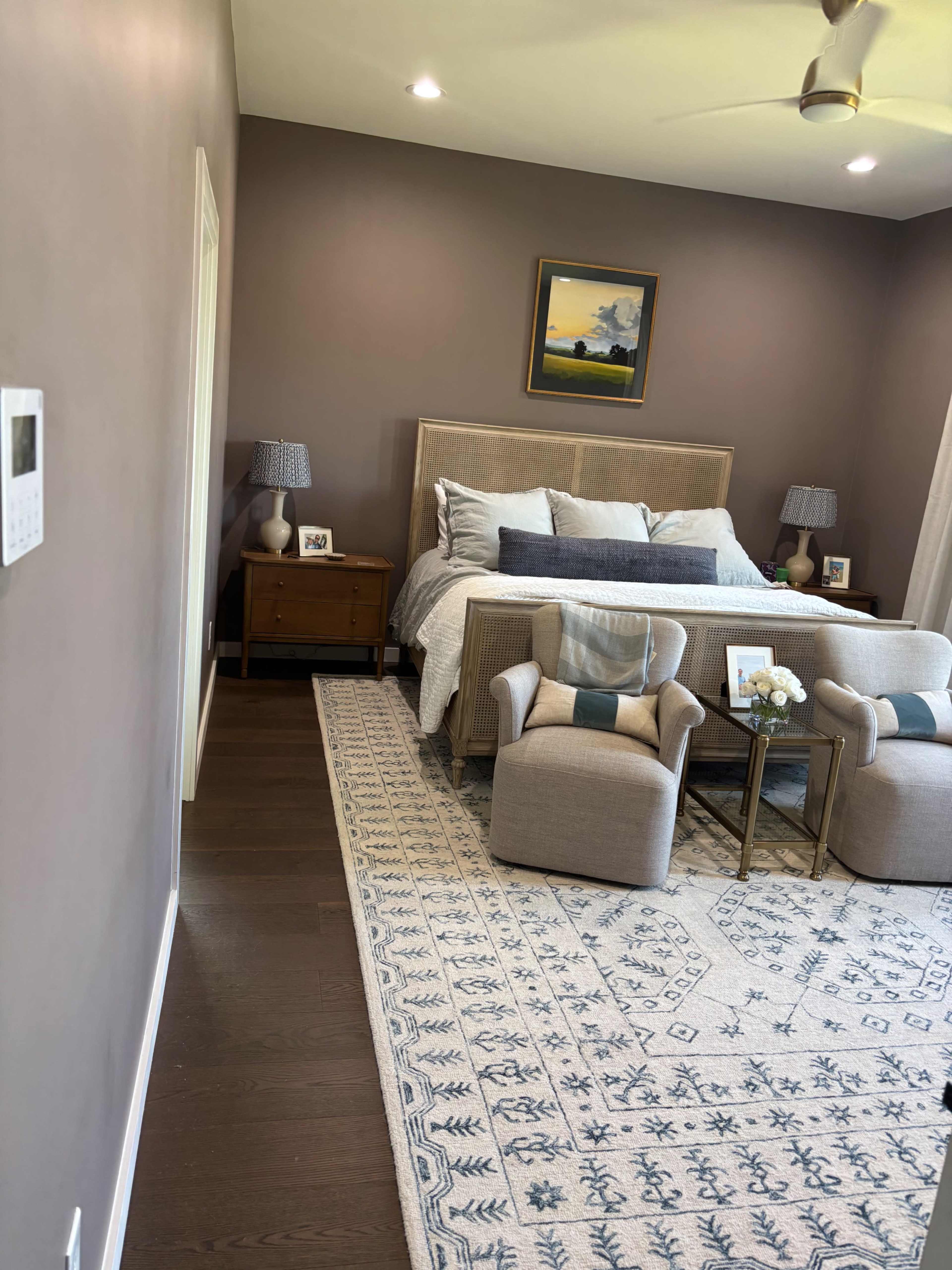 The image shows a modern bedroom with a bed, two upholstered chairs, a nightstand, and a decorative area rug.