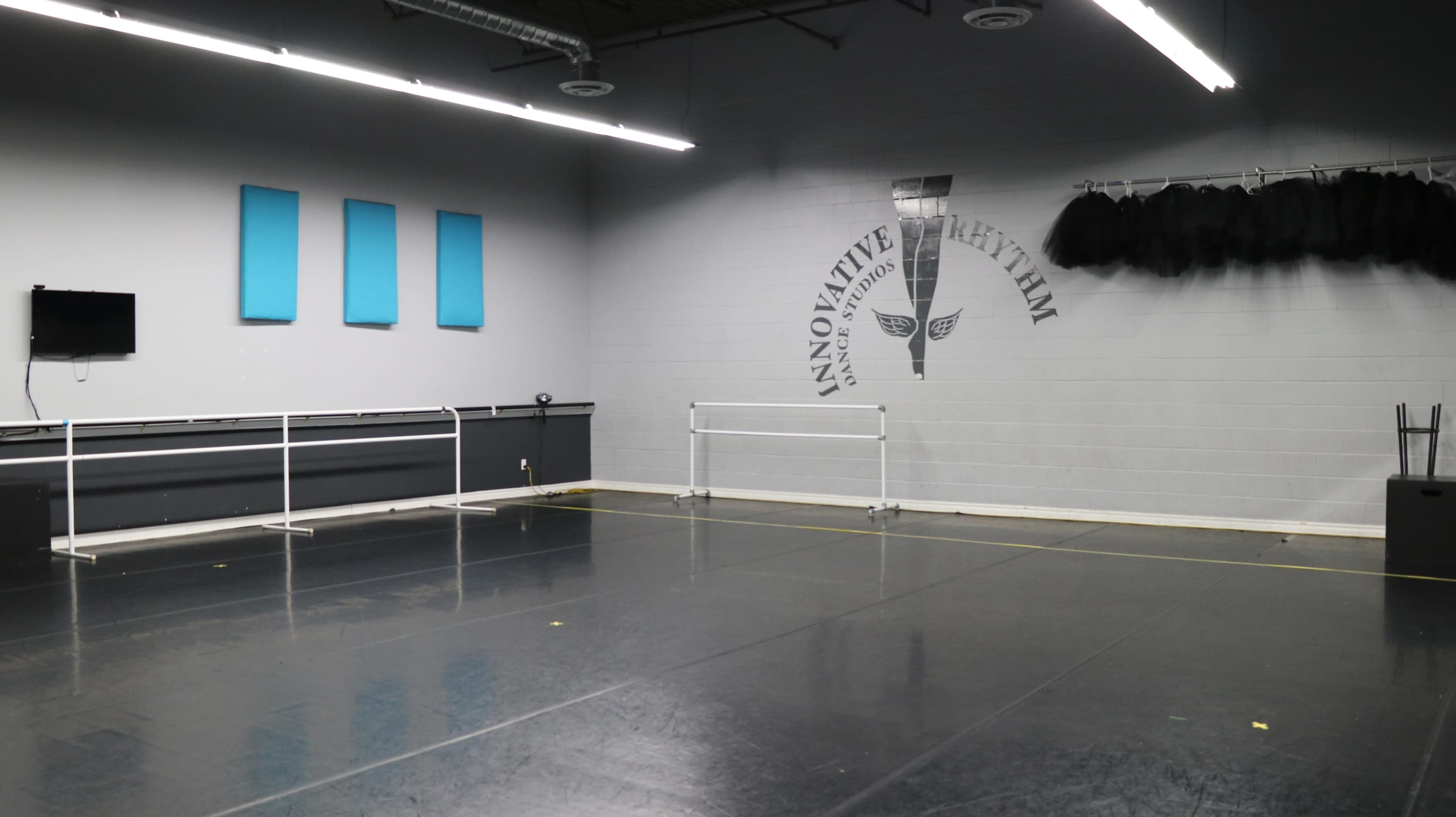 The image shows a spacious dance studio with a mirrored wall, ballet barres, and gray walls featuring blue panels and the words "Innovative Rhythm."