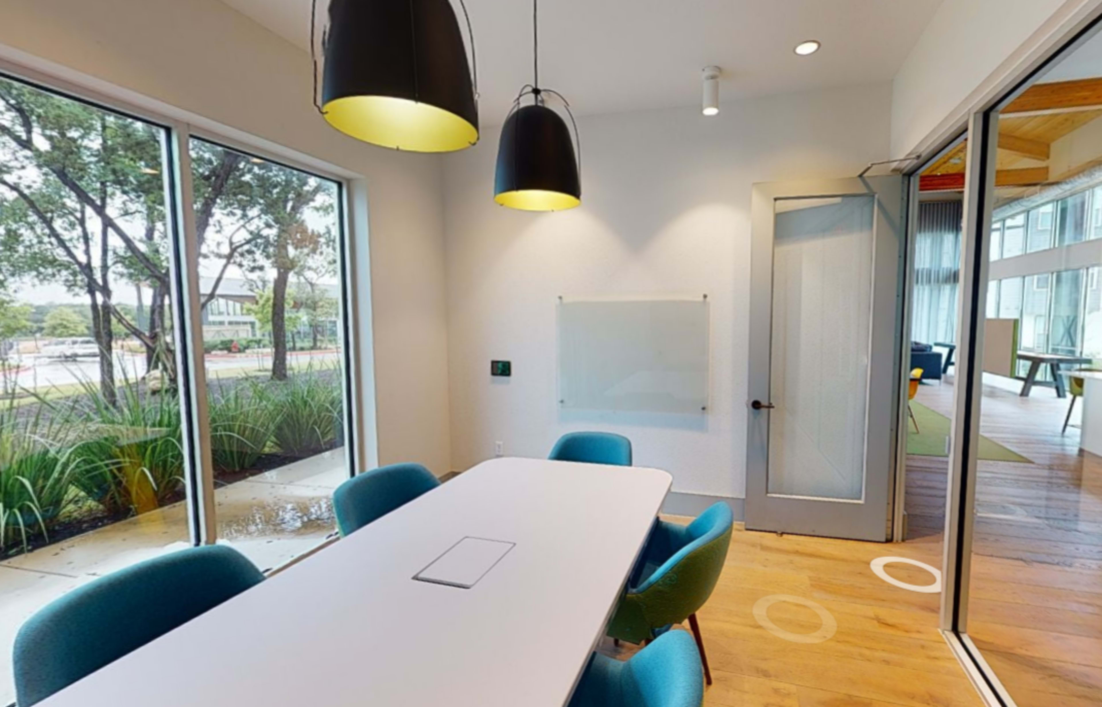 A modern conference room features a long white table surrounded by blue chairs, with large windows offering views of greenery outside.