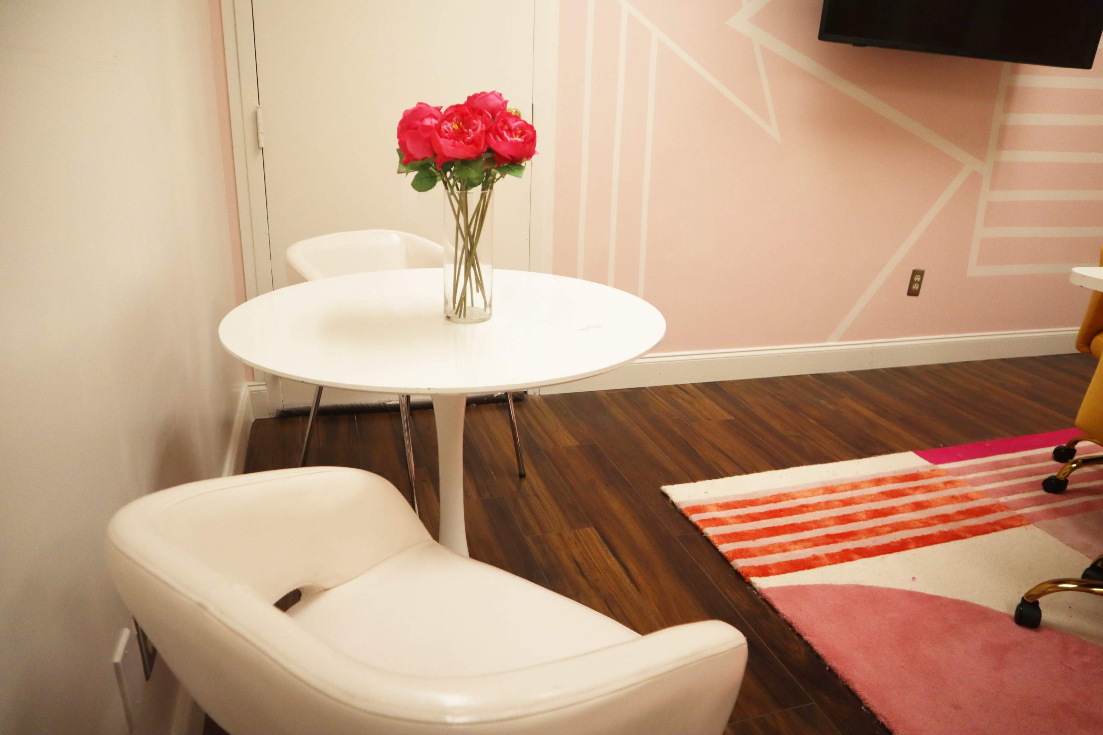 A white table with a vase of pink flowers is positioned next to a white chair in a room with pink geometric wall designs and a colorful rug.