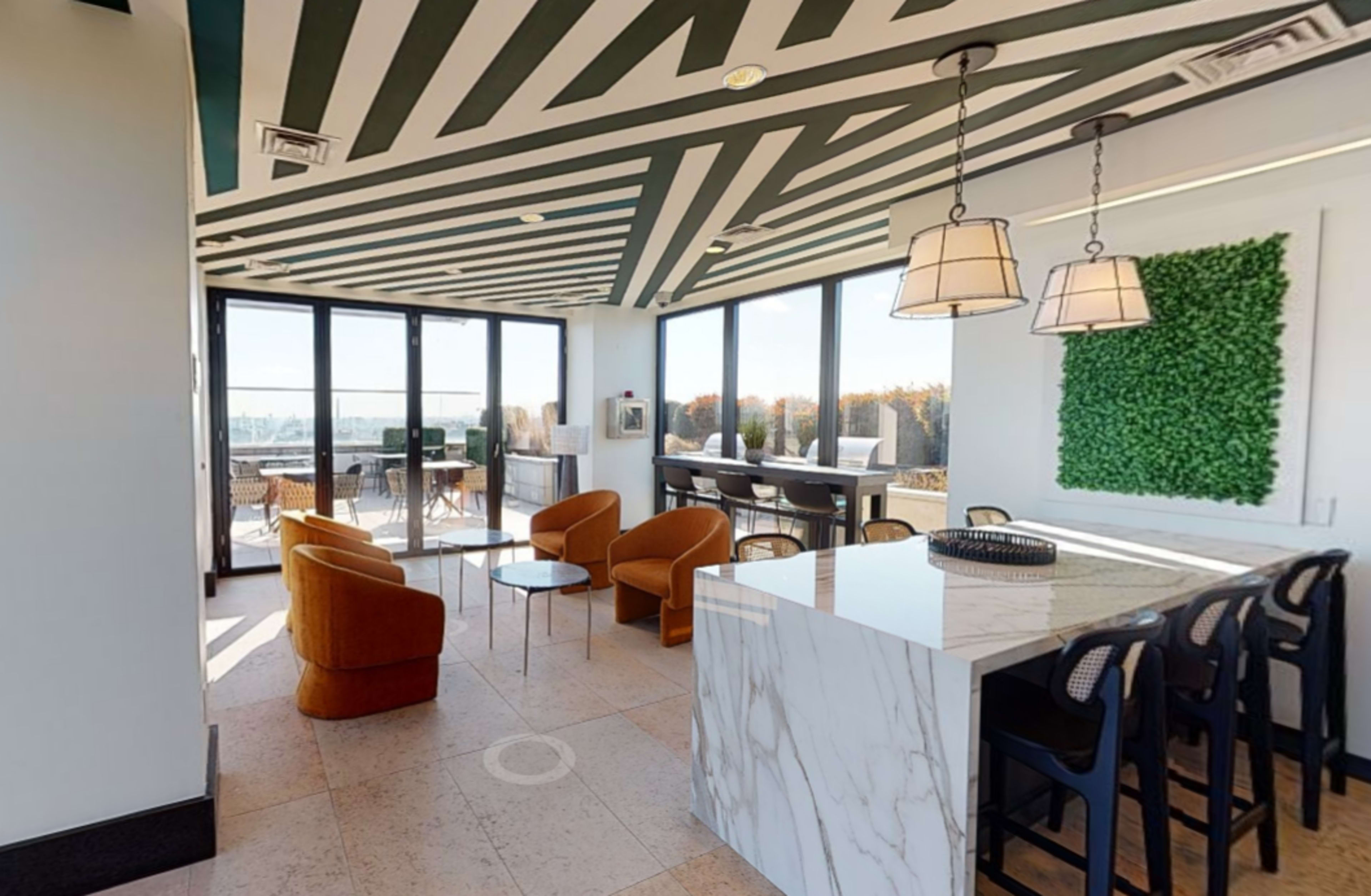 A modern indoor lounge area with striped ceilings, large windows, and a marble countertop, furnished with orange chairs and a green wall feature.