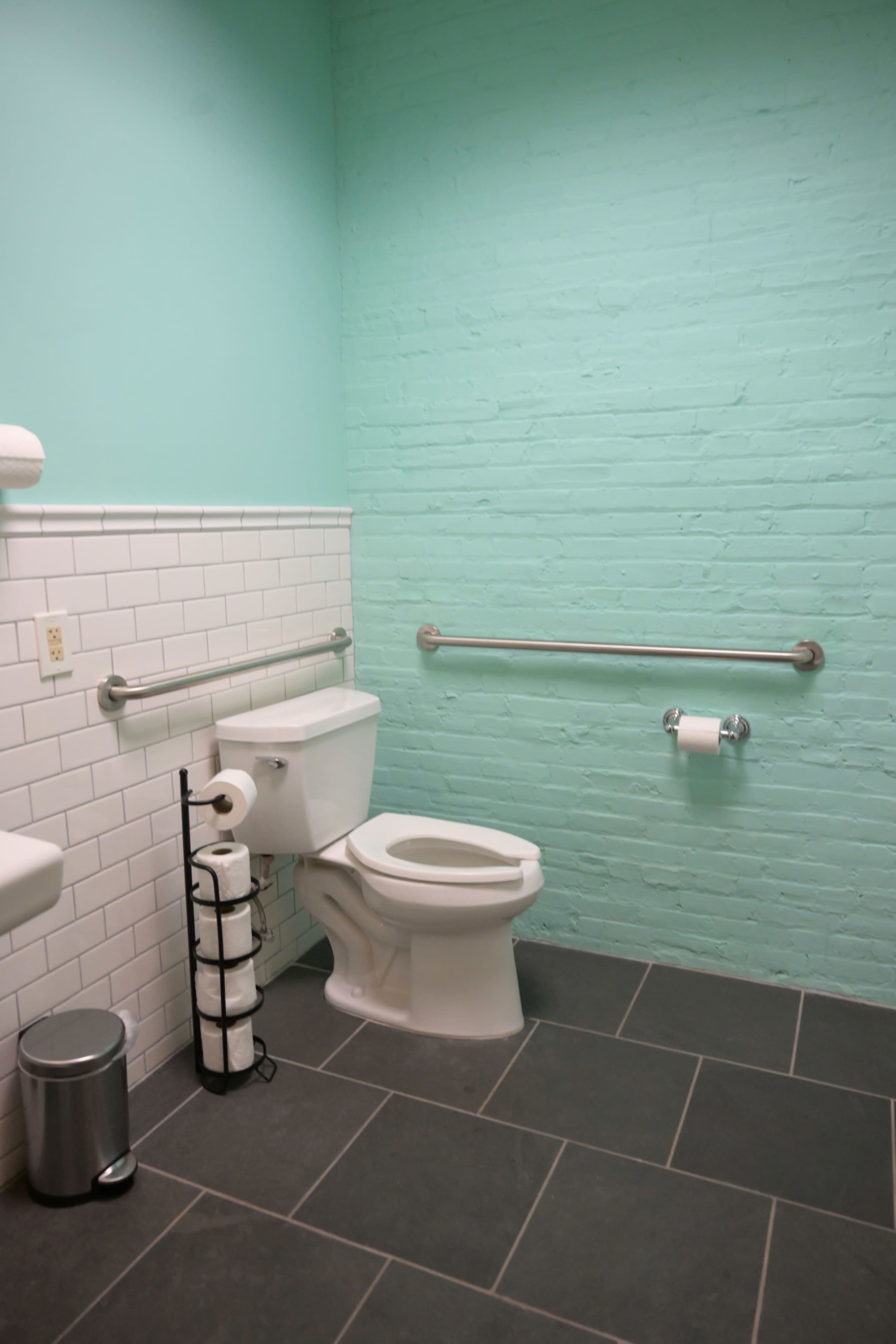 The image shows a bathroom with a turquoise brick wall, white tiled accents, a toilet, a sink, and gray flooring.