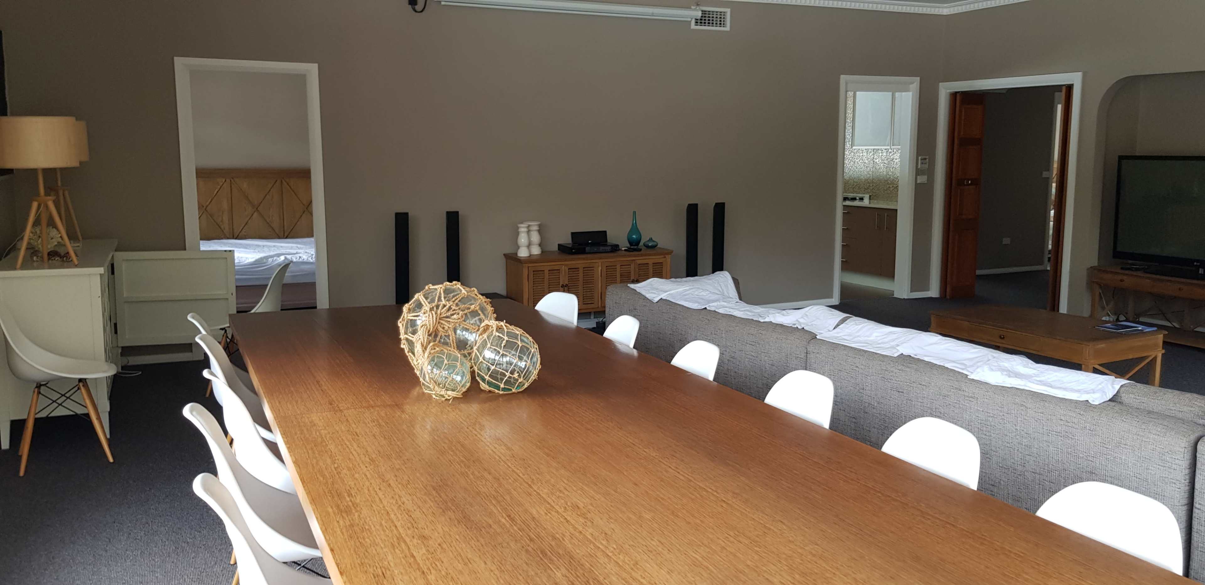 A spacious living area with a long wooden dining table, white chairs, a gray sofa, and a coffee table, all set in a room with light walls and dark carpet.
