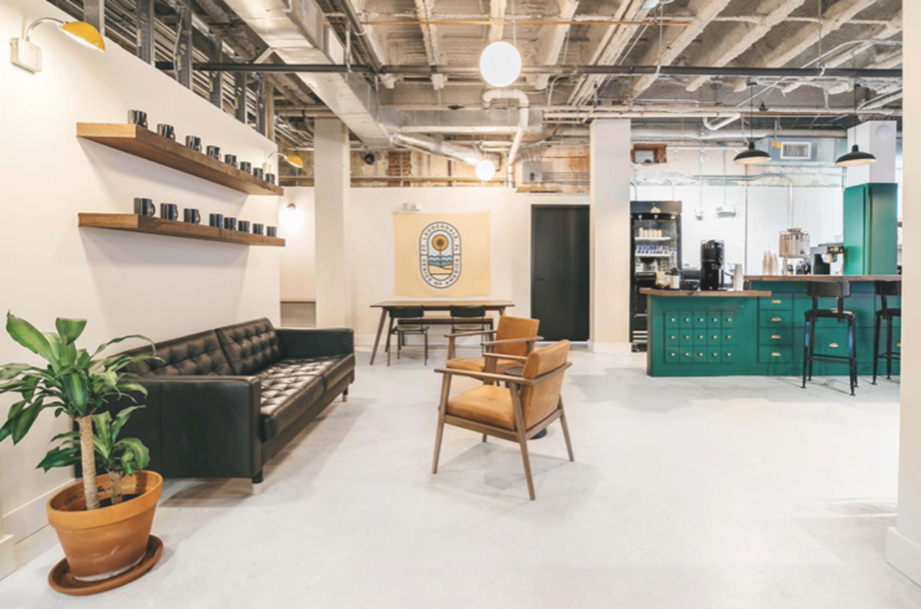 A modern office space featuring a black leather couch, wooden shelves with mugs, a green kitchen area, and a brown armchair.