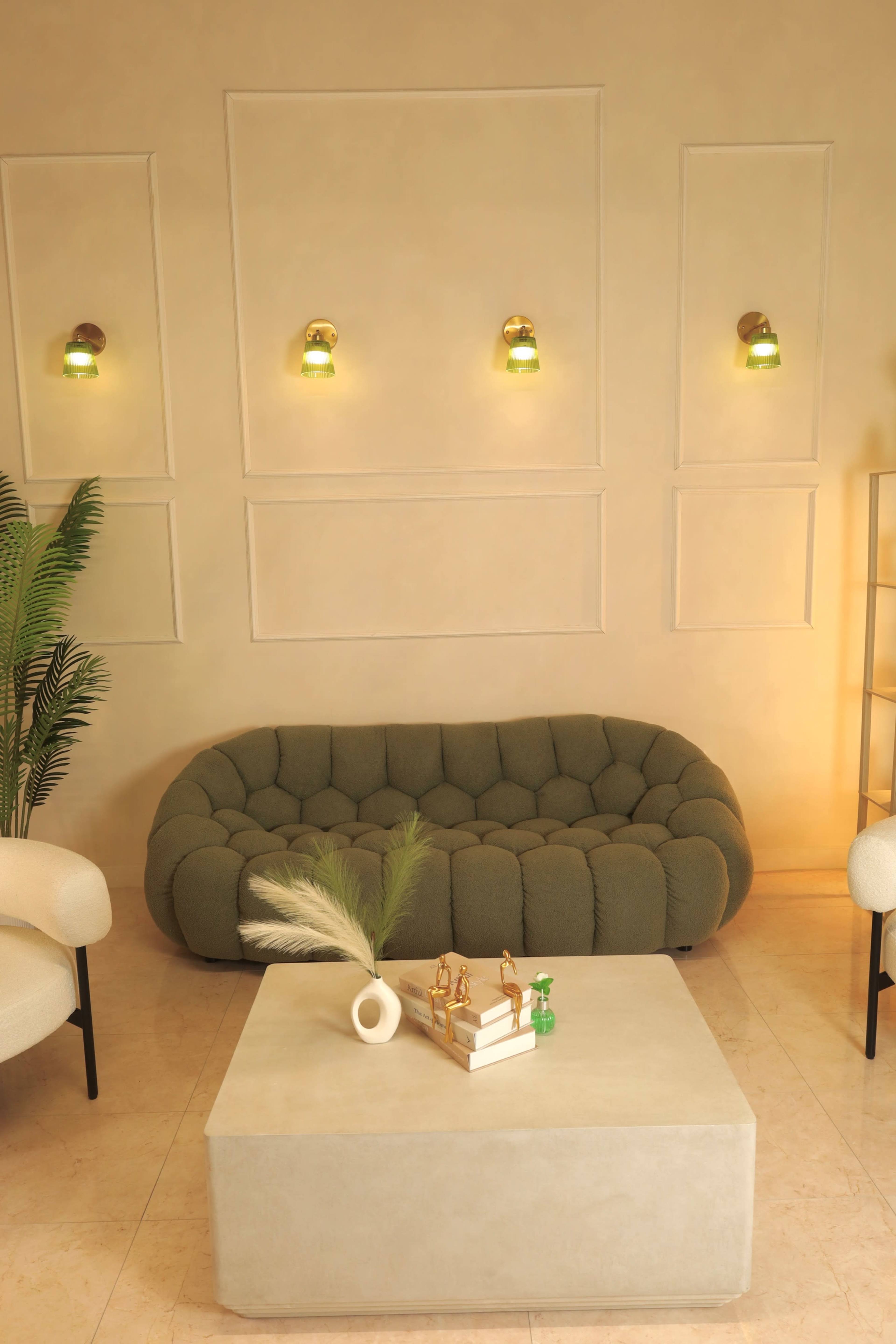 A modern living room features a green tufted sofa, a light-colored coffee table with decorative items, and wall-mounted lamps.
