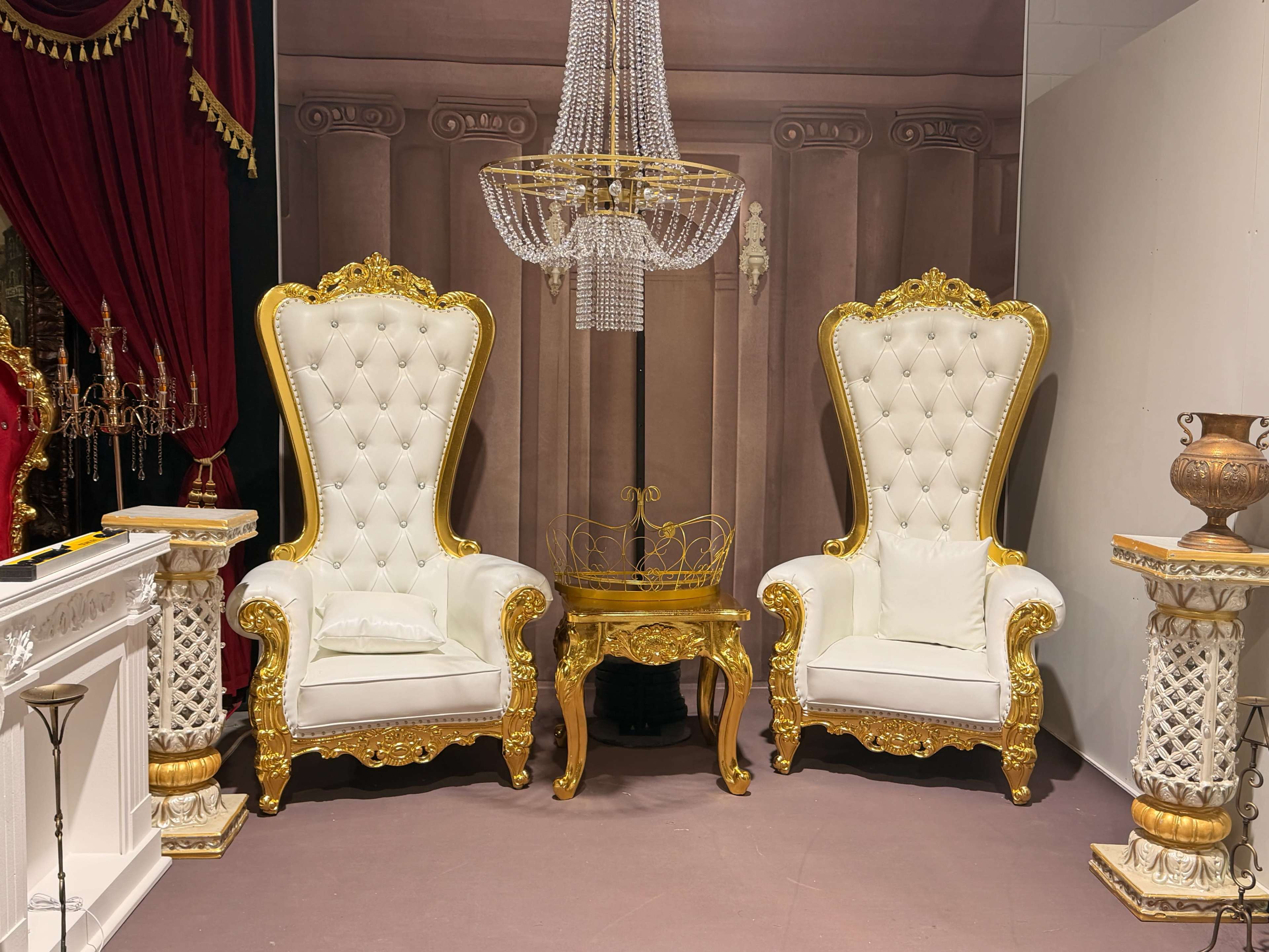 The image shows two ornate white and gold armchairs flanking a small round table, set against a backdrop featuring columns and a chandelier.