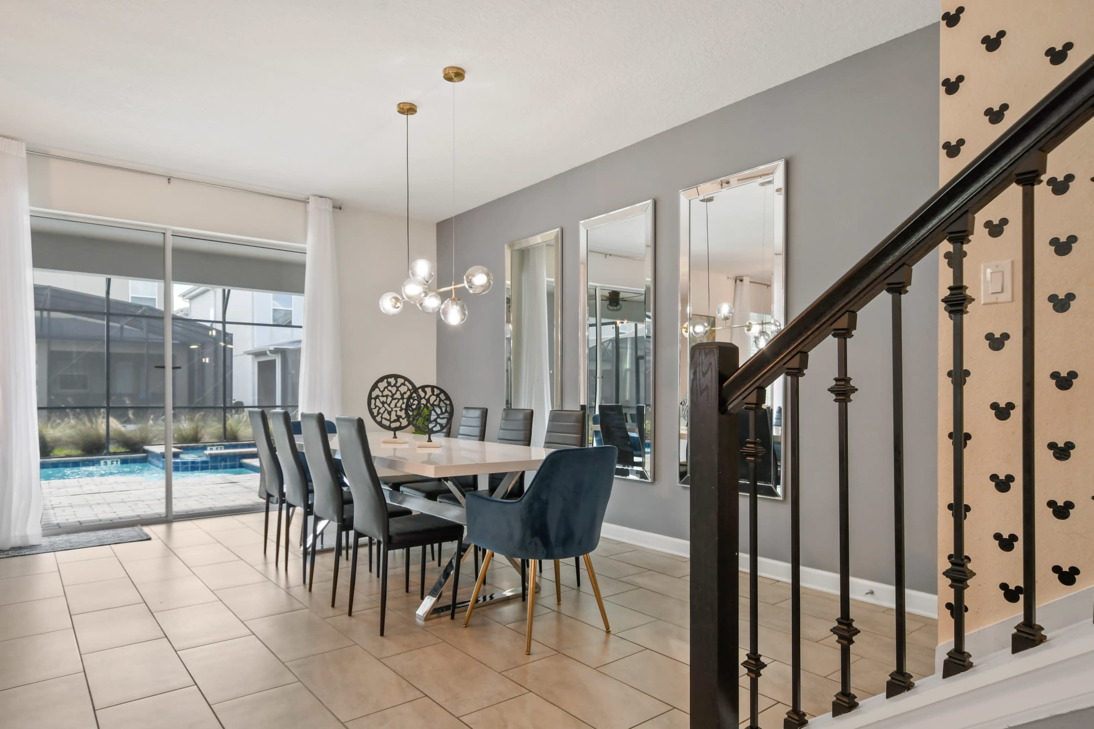 A modern dining area with a long table and six chairs, large mirrors, and a staircase, adjacent to a pool view through sliding glass doors.