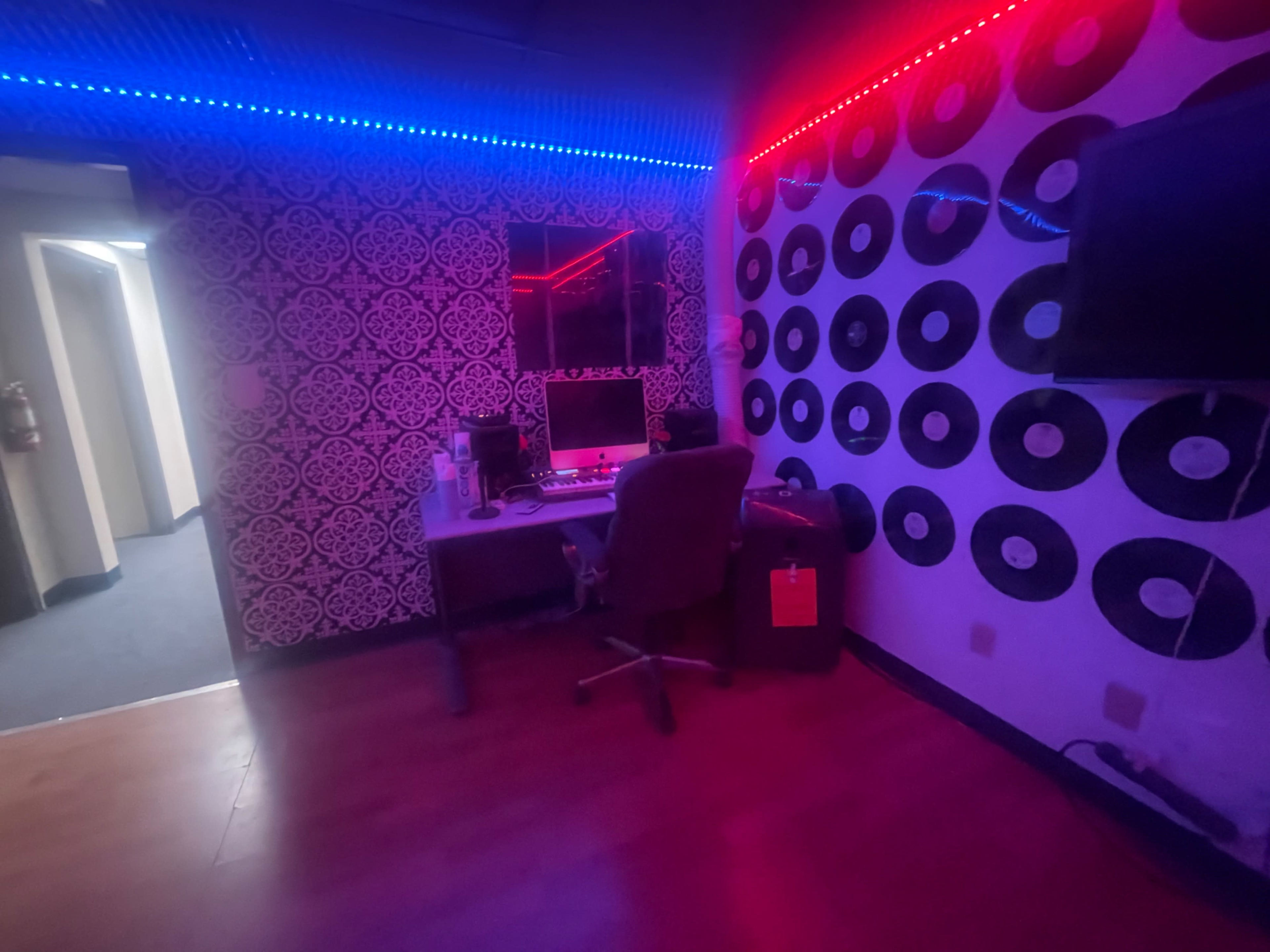 A music recording studio with a desk, computer equipment, and walls decorated with vinyl records, illuminated by red and blue LED lights.