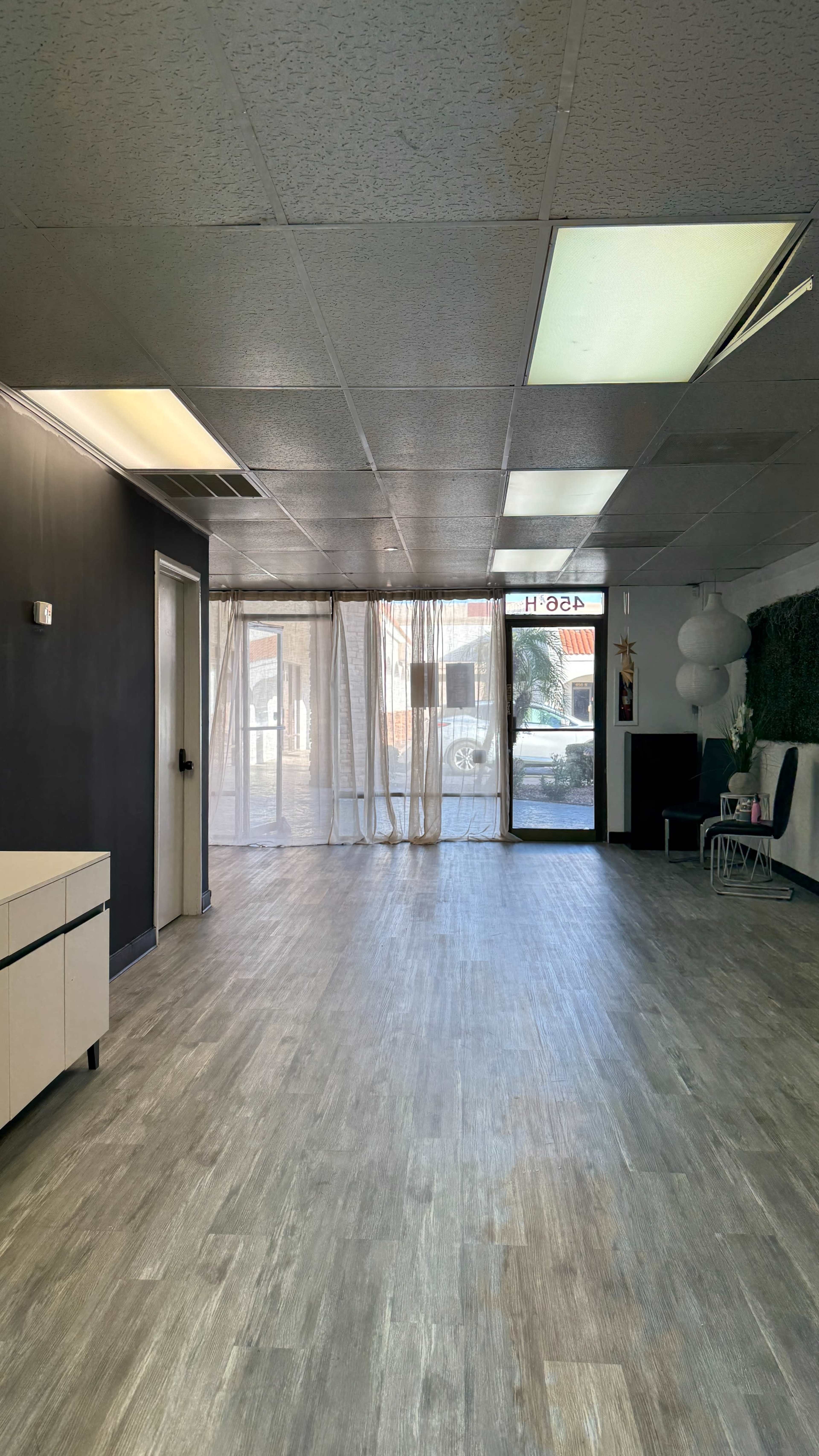 The image shows a spacious, mostly empty room with a wood-like floor, light-colored walls, and large windows covered with sheer curtains.
