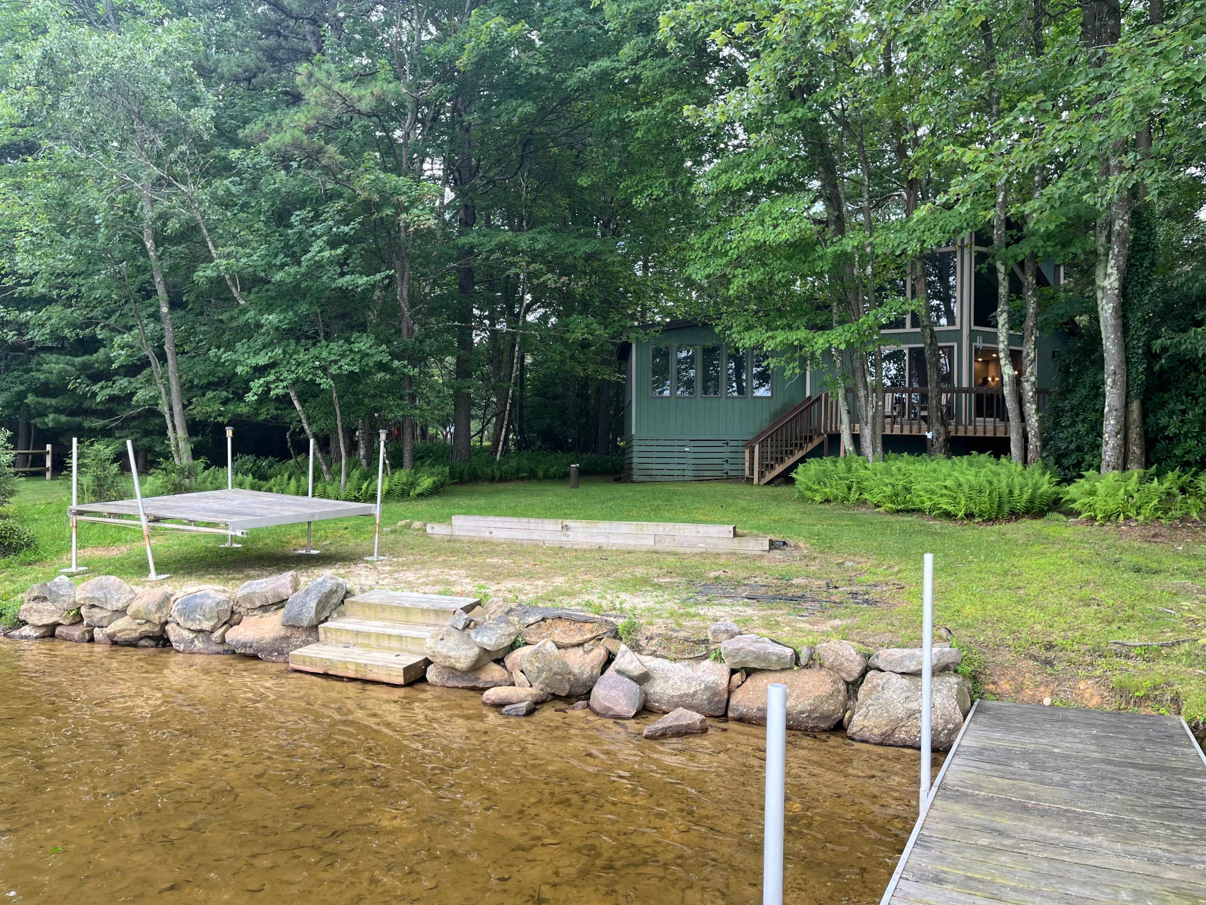 A grassy area with a wooden dock leads to a calm lake in front of a green cabin partially obscured by trees.