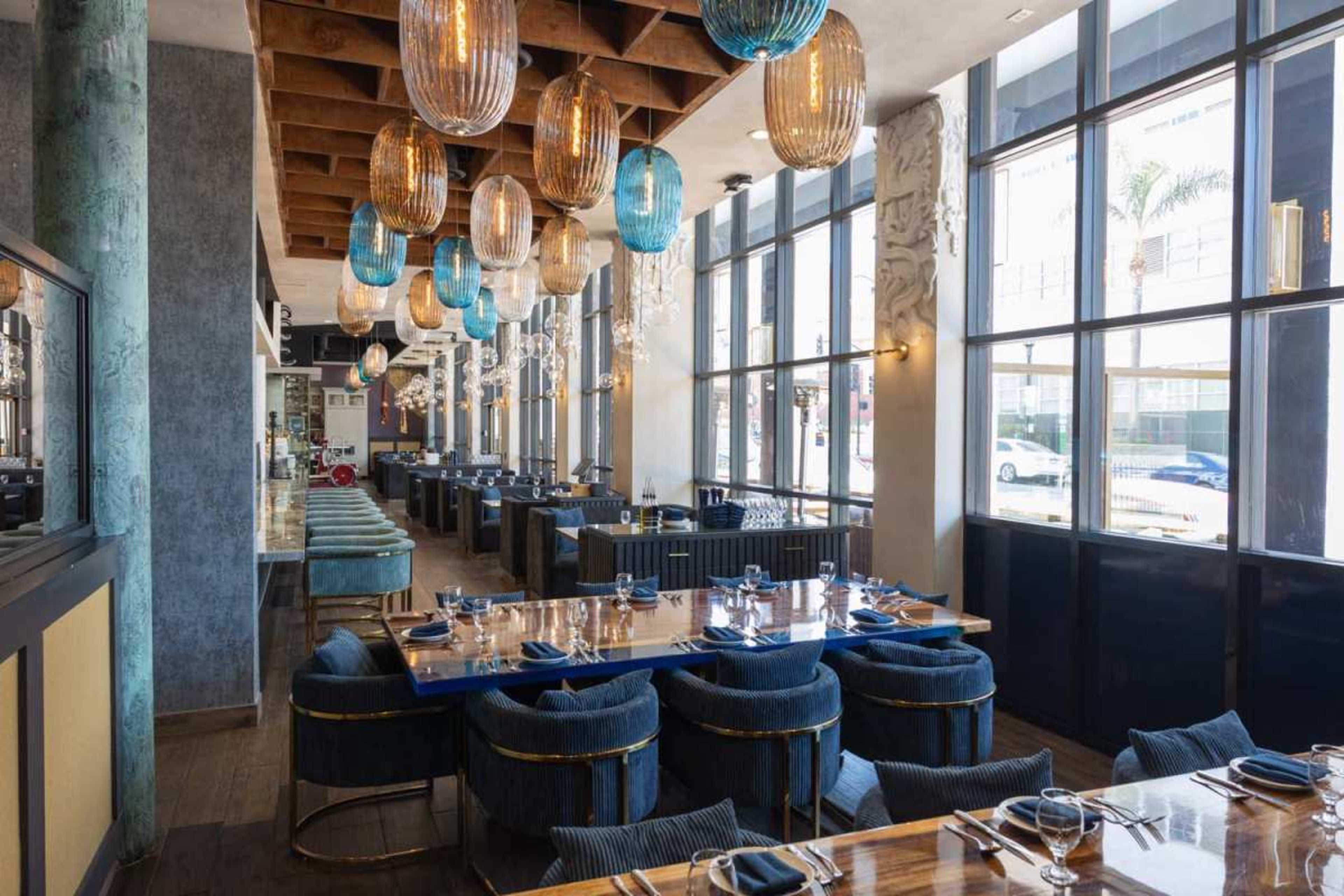 The image shows a modern restaurant interior featuring blue and gold decor, large windows, and illuminated pendant lights hanging from the ceiling.