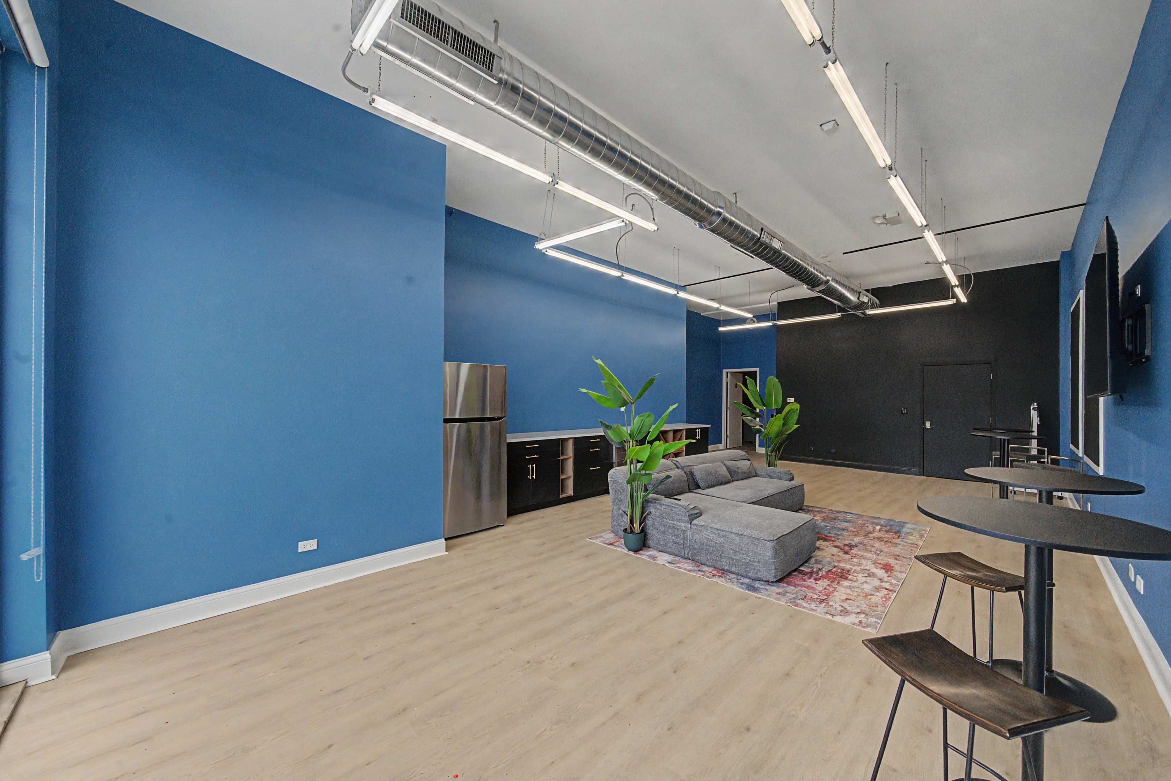 The image shows a spacious room with blue walls, featuring a gray sofa, a kitchenette with a refrigerator, a large black wall, and a couple of standing tables.