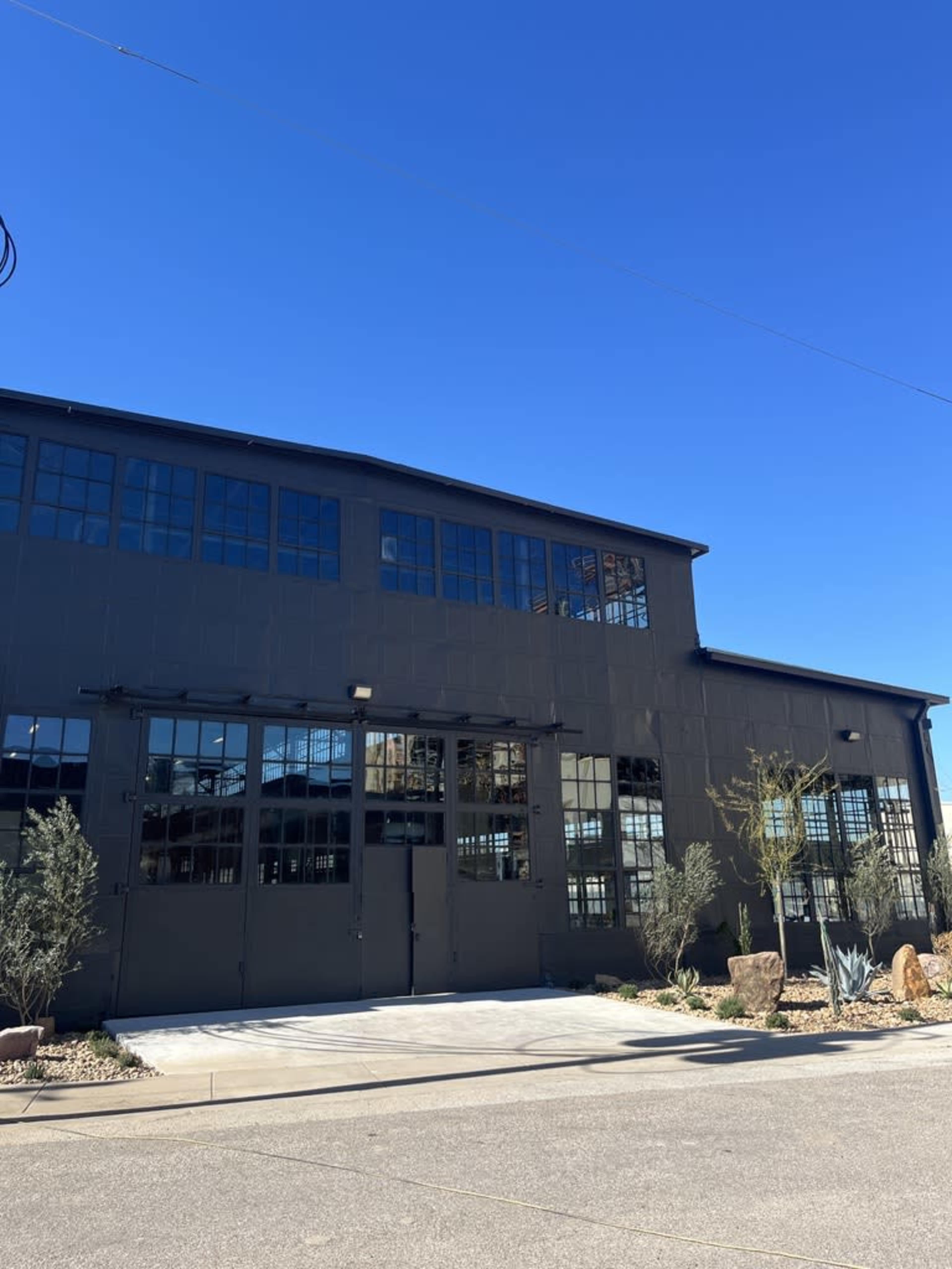The image shows a large, modern industrial building with large windows, a black exterior, and a landscaped area in front featuring rocks and plants under a clear blue sky.