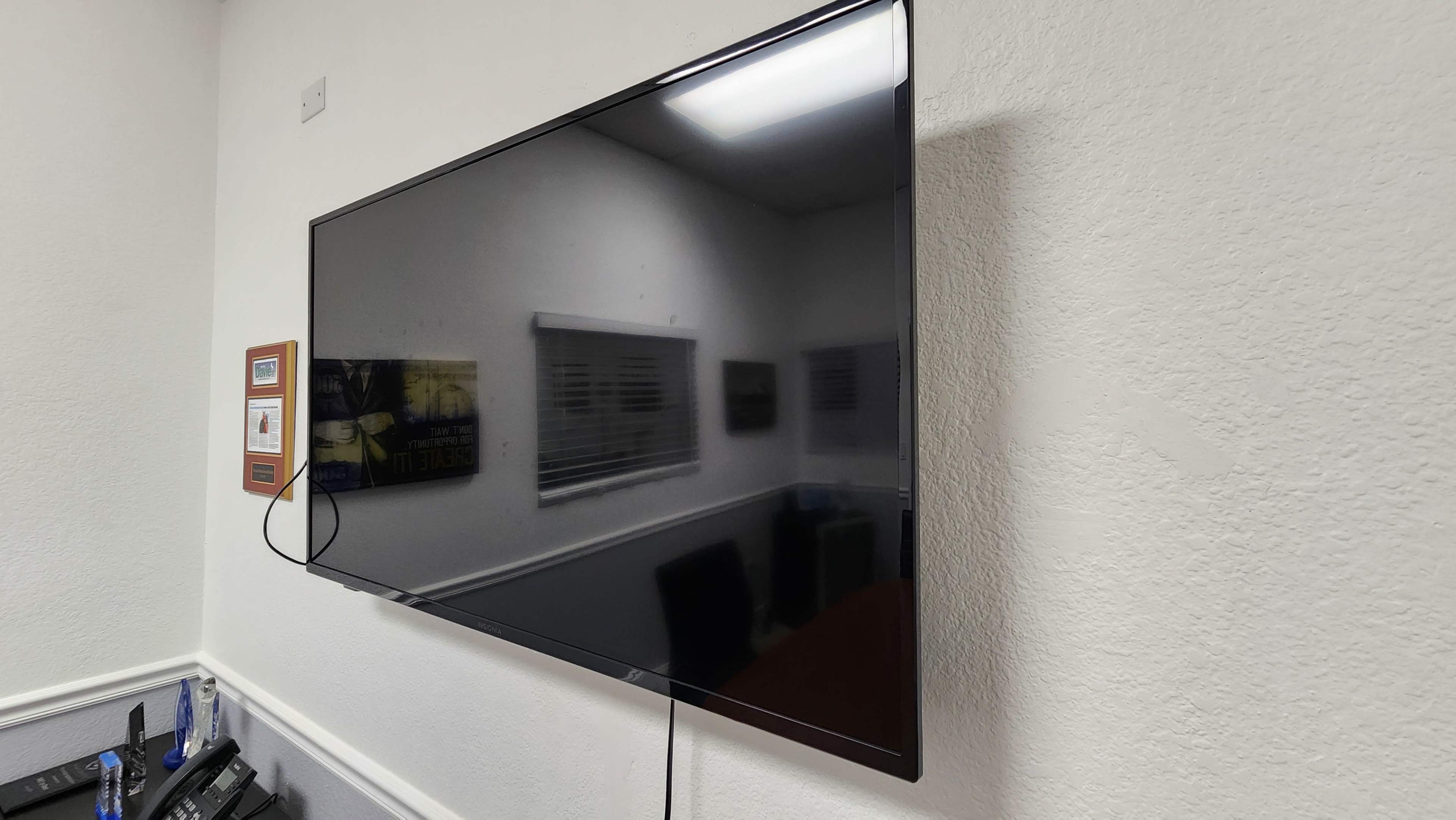 A flat-screen television is mounted on a white wall in a room with a conference table and windows in the background.