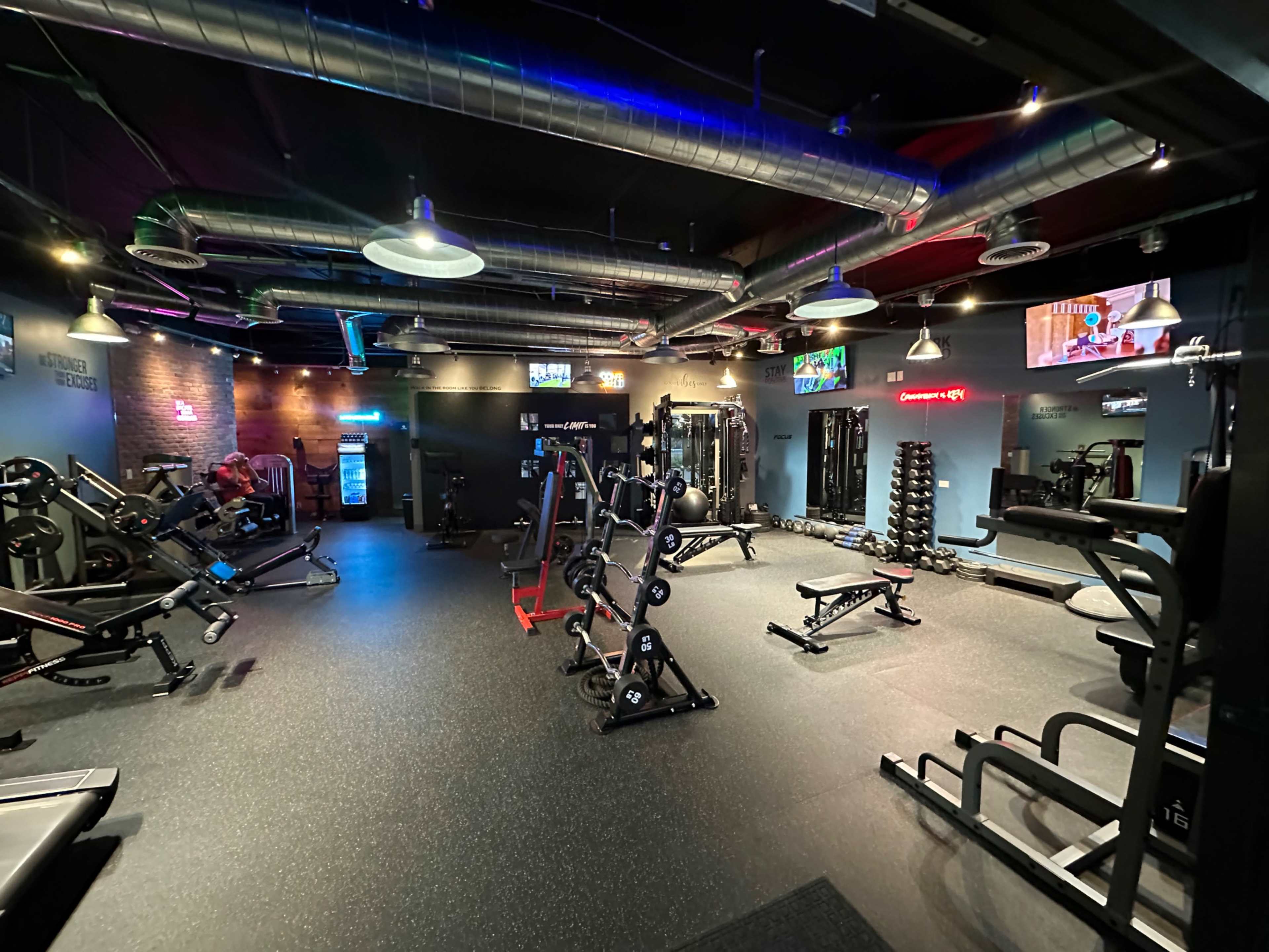 The image shows a modern gym interior with various fitness equipment arranged around the space, including weight benches, machines, and free weights, under bright lighting.