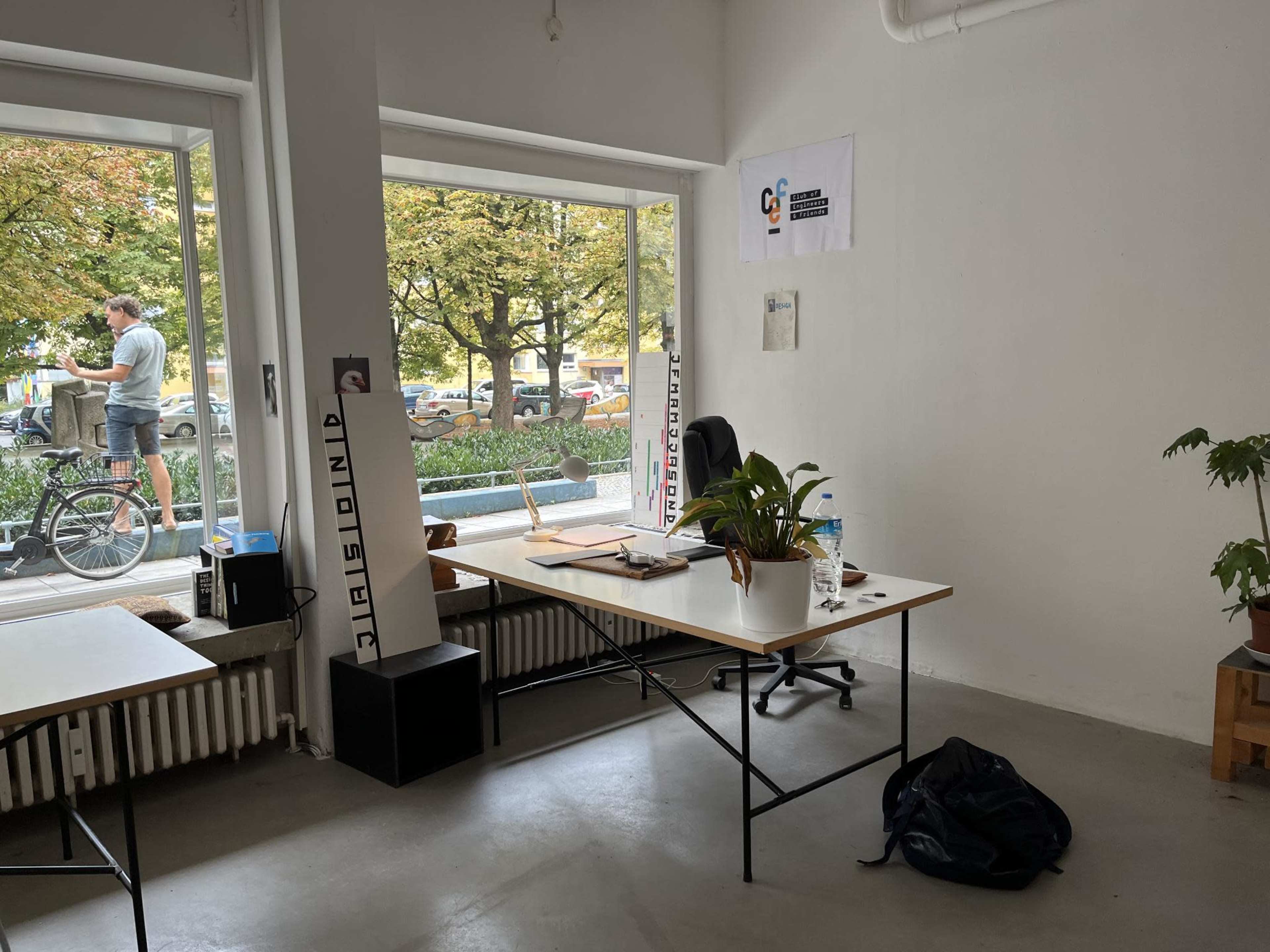 An office space features a desk with a plant, a chair, and a backdrop of large windows showing a person outside with a bicycle.