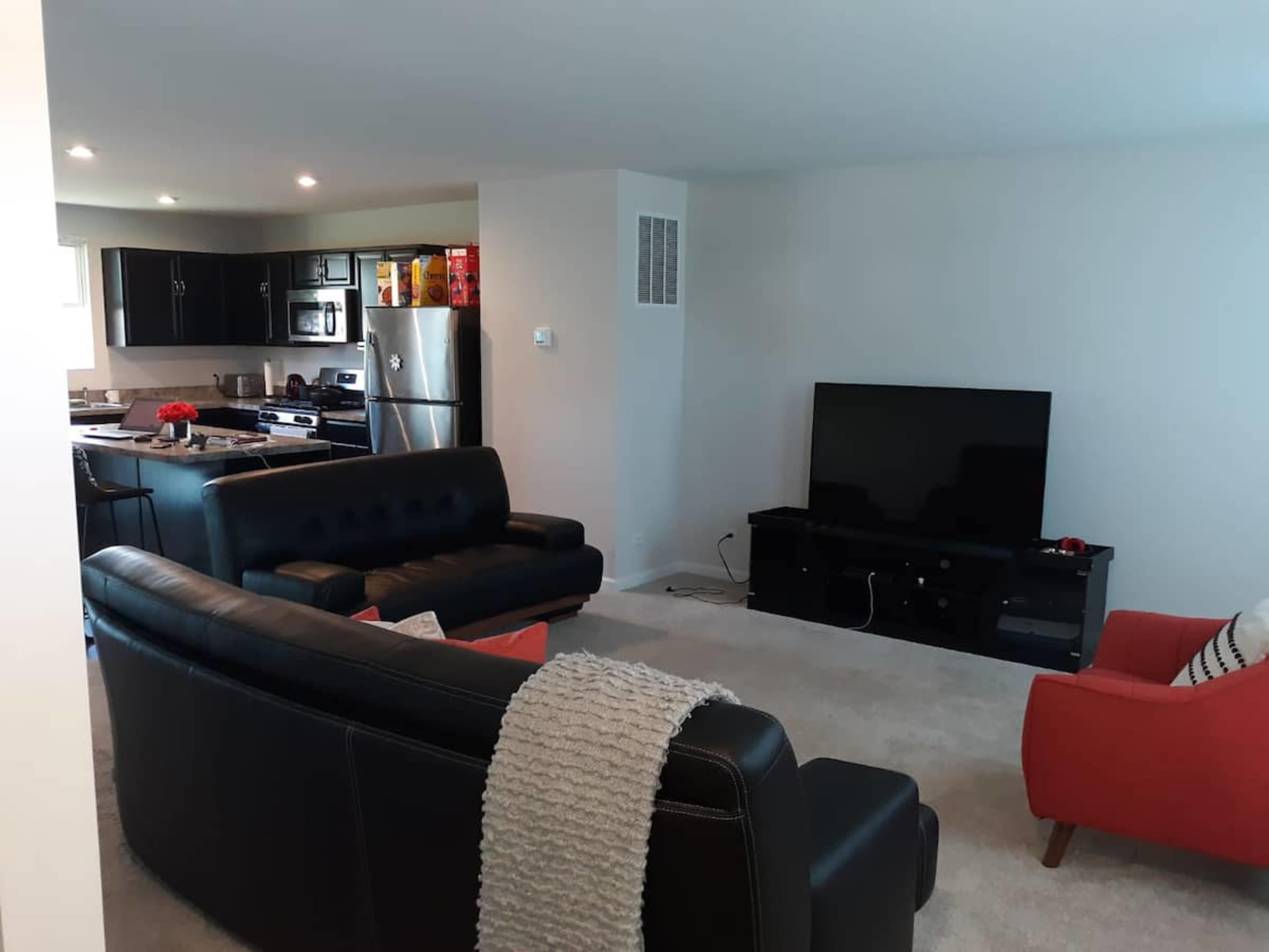 A modern living room with two black sofas, an orange armchair, a television on a stand, and an open kitchen area in the background.