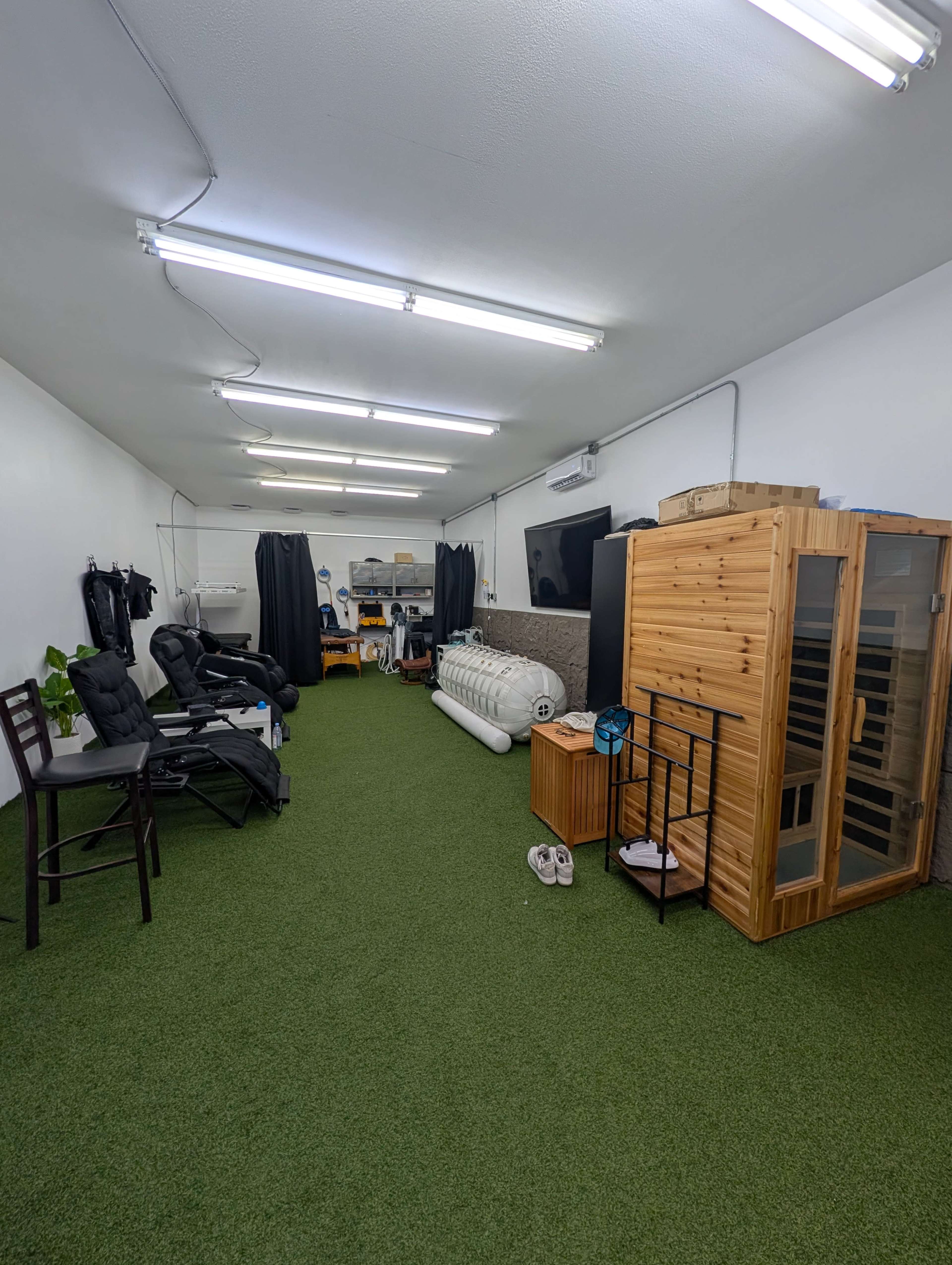 The image shows a spacious room with artificial grass flooring, featuring seating areas, exercise equipment, and a wooden sauna situated against the wall.
