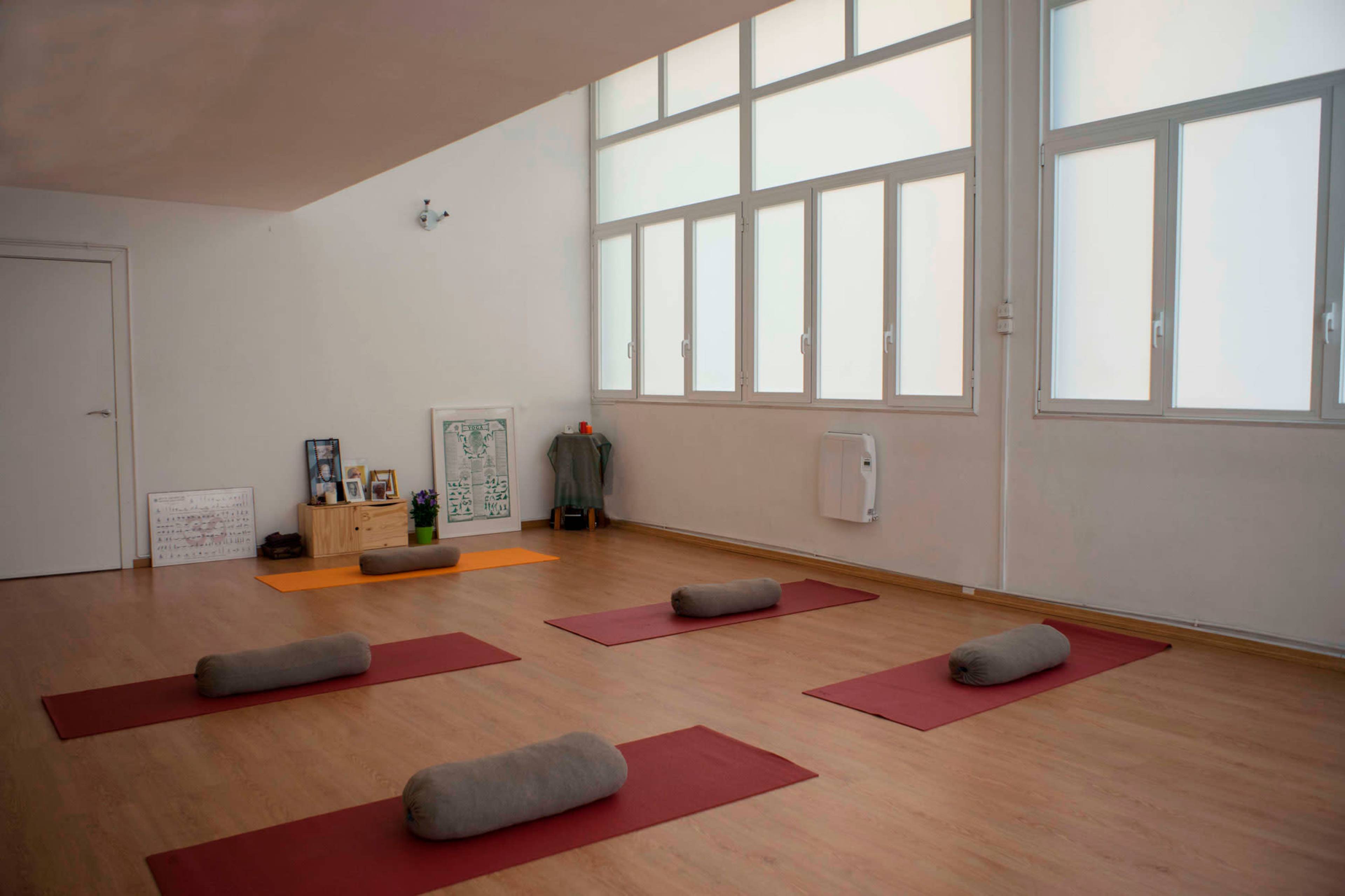 A spacious room with wooden floors, featuring several yoga mats arranged on the floor and large windows allowing natural light to fill the space.